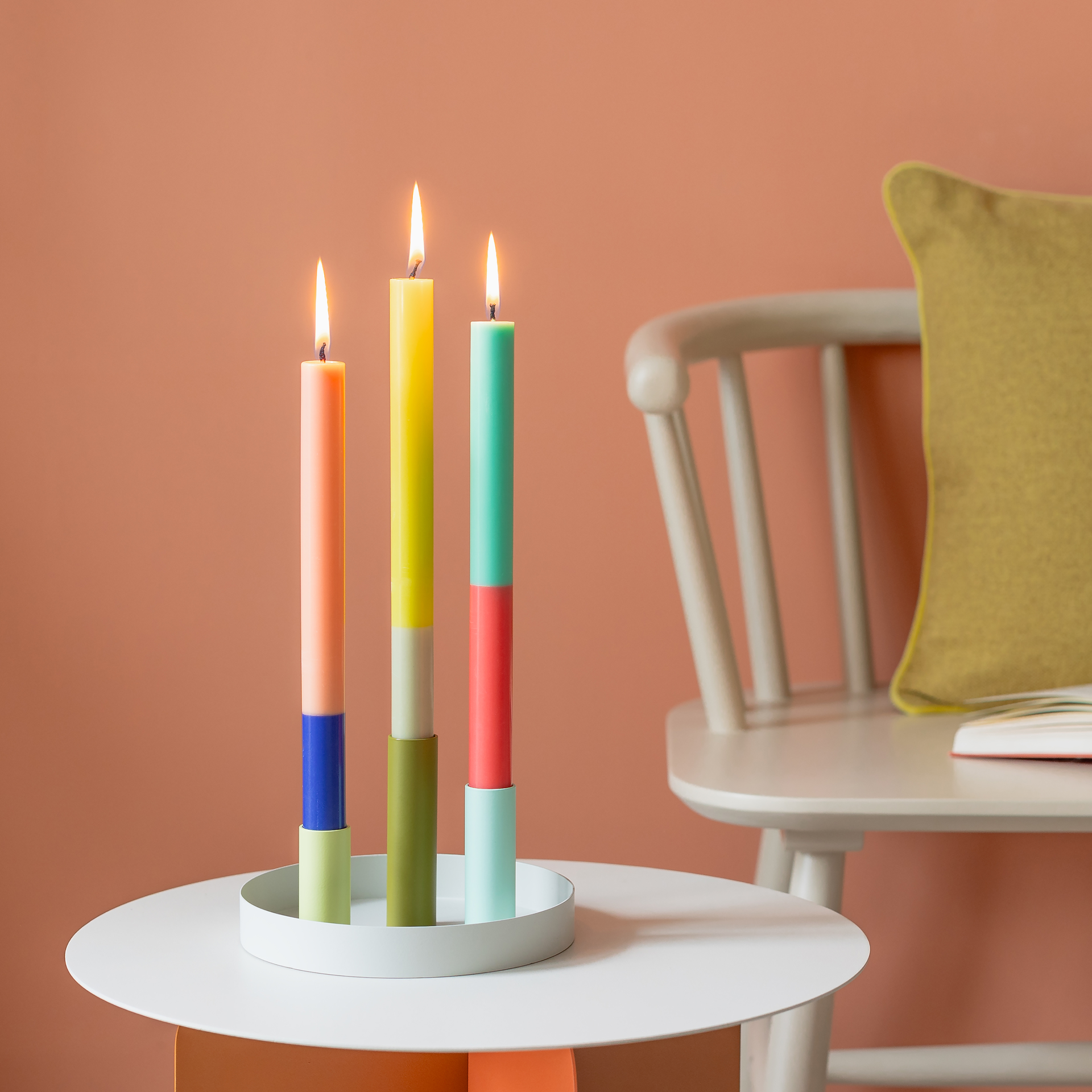 Drei bunte Stabkerzen des 'Ibiza' Sets, jede mit zwei Farbblöcken, stehen auf einem weißen Tisch. Die Kerzen sind angezündet. Im Hintergrund ein Stuhl.