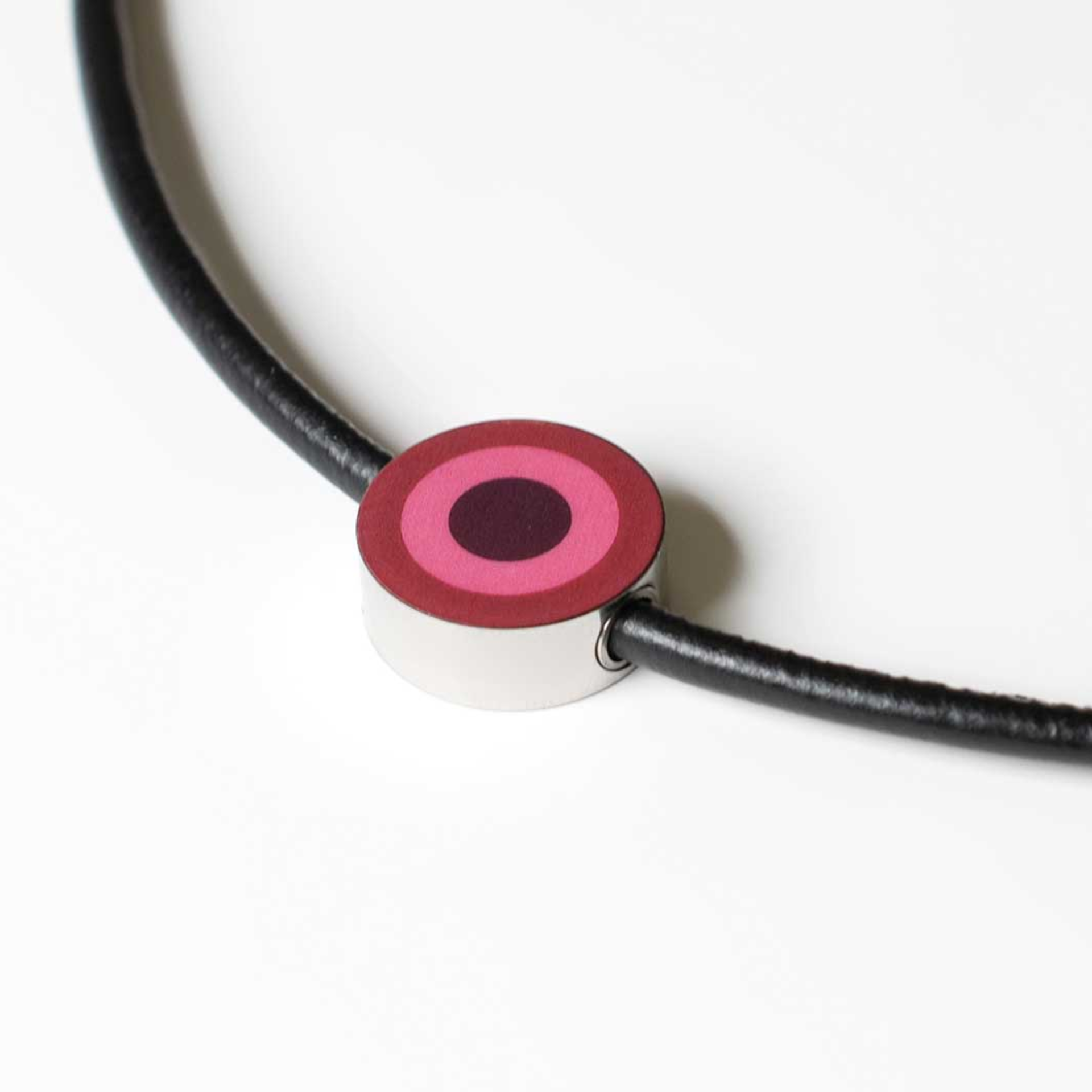 Ciondolo gioiello: gioiello rotondo con cerchi concentrici in rosa e rosso scuro su un nastro nero. Il ciondolo è in argento lucido.