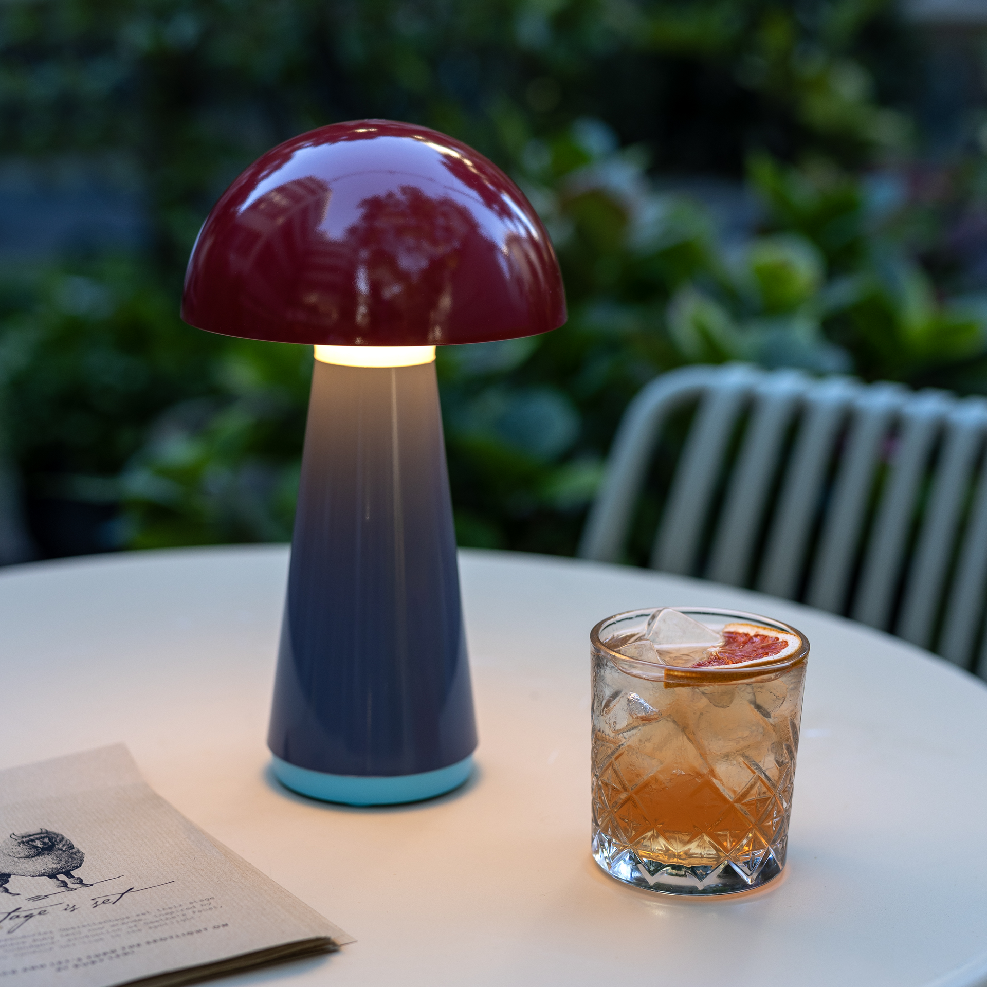 Eine rote Tischleuchte 'Bob' im Pilzdesign leuchtet neben einem Cocktailglas mit Eiswürfeln auf einem Tisch im Freien. Im Hintergrund sind grüne Pflanzen sichtbar.