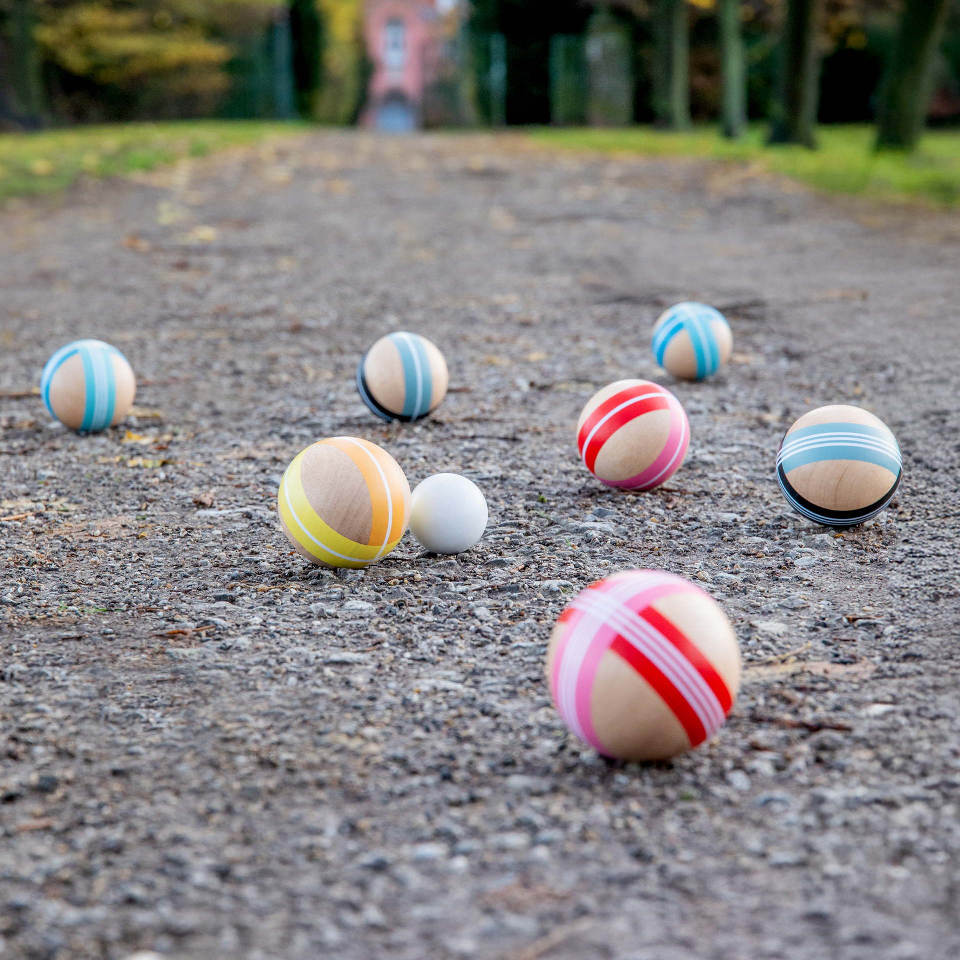 Boccia-Set: Acht große Holzkugeln mit Streifenmuster liegen auf einem Kiesweg, neben einer kleinen weißen Zielkugel.