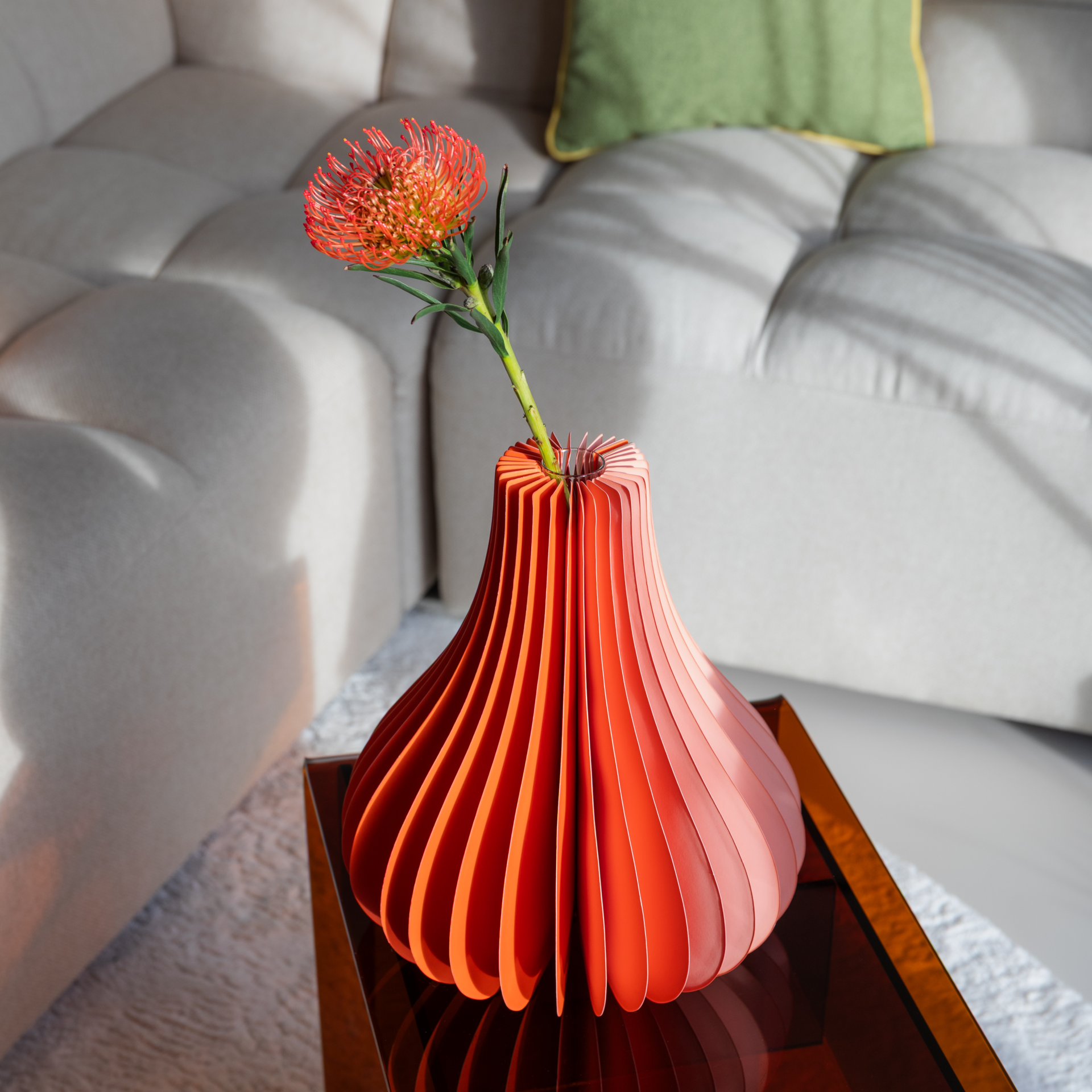 Un vase orange à côtes contenant une fleur de protéa est posé sur une table basse en verre devant un canapé clair.