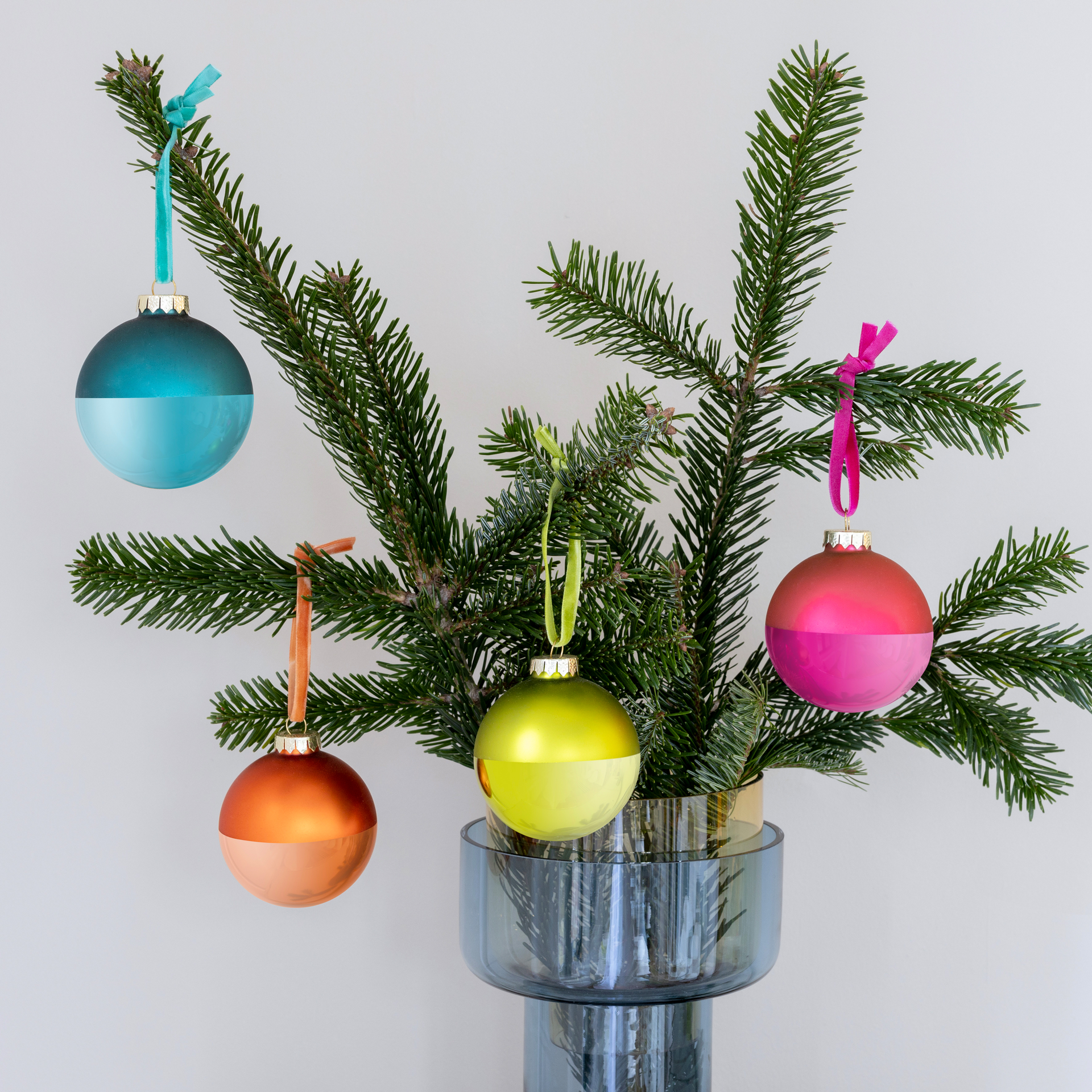 Detailaufnahme von vier zweifarbigen Weihnachtskugeln am Tannenzweig in einer Vase, inklusive Samtbändern zur Aufhängung. Die Farben sind blau, orange, grün und pink.
