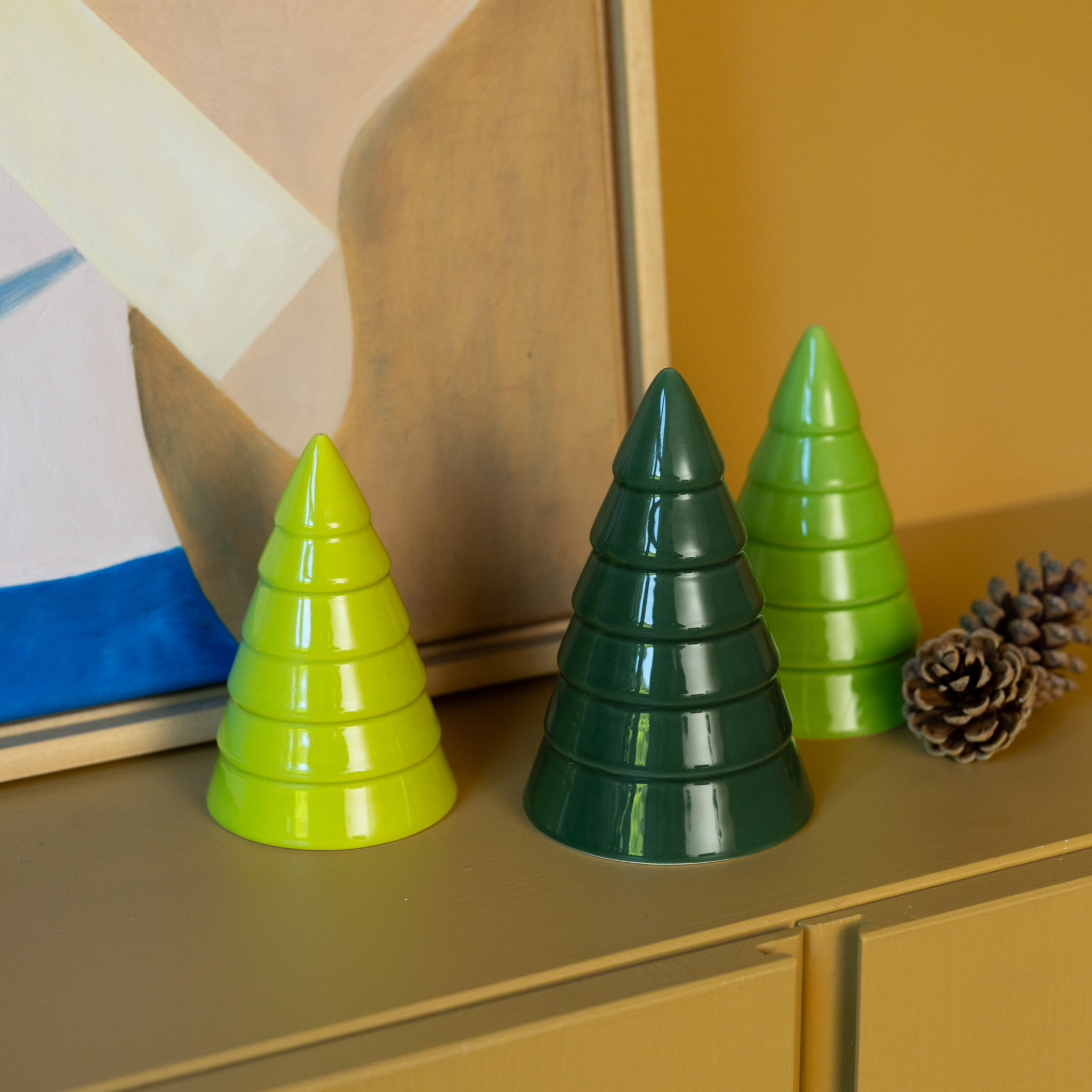 Tre alberi decorativi in ceramica a forma di abete su sfondo bianco. Da sinistra a destra: verde scuro, verde medio, verde chiaro. Smaltati lucidi con scanalature orizzontali che riproducono rami stilizzati e una texture a strati.
