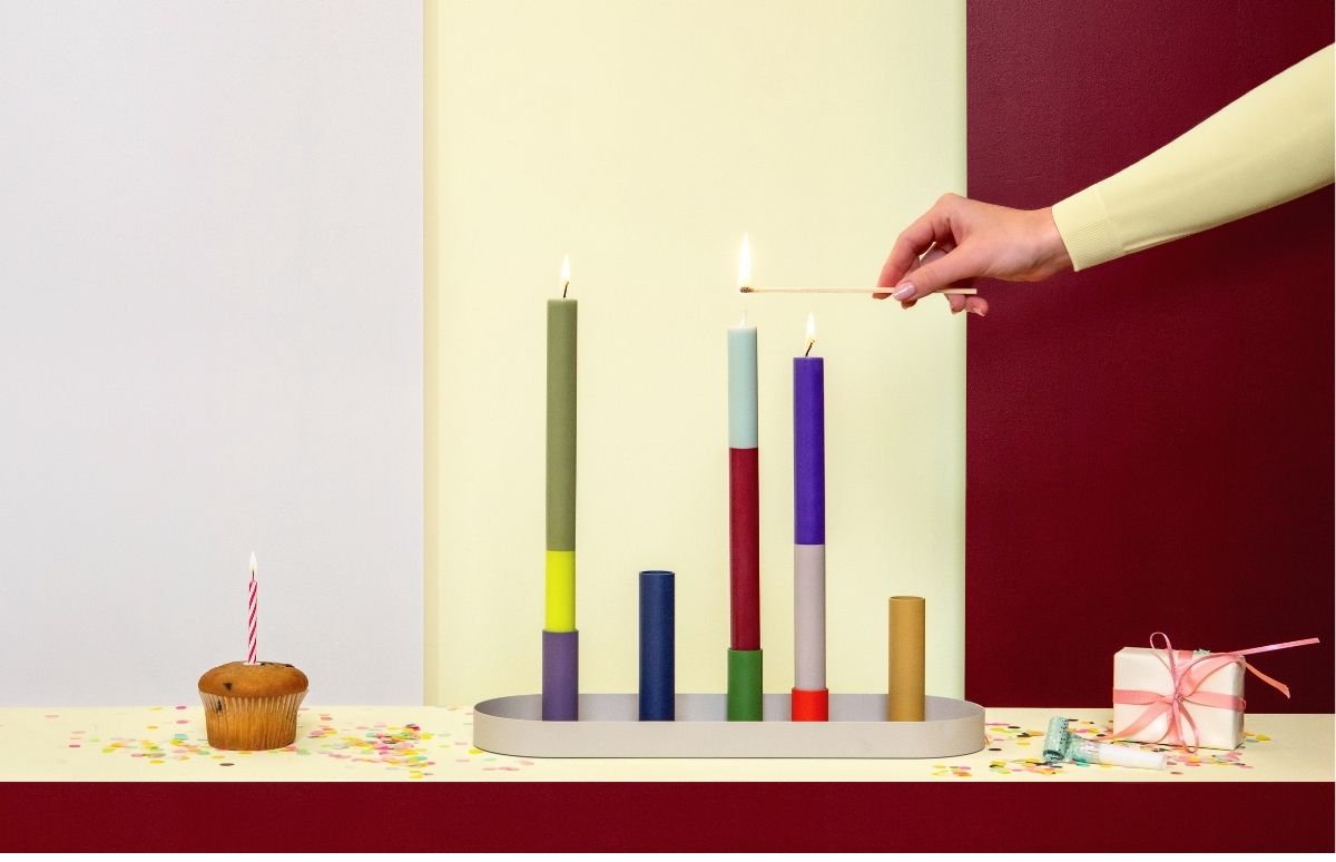 Eine Hand zündet eine mintgrüne Kerze auf einem ovalen Tablett an, mit weiteren bunten Kerzen in verschiedenen Höhen. Links ein Cupcake mit gestreifter Geburtstagskerze. Rechts Geschenk mit rosa Band und Konfetti auf gelber Fläche.