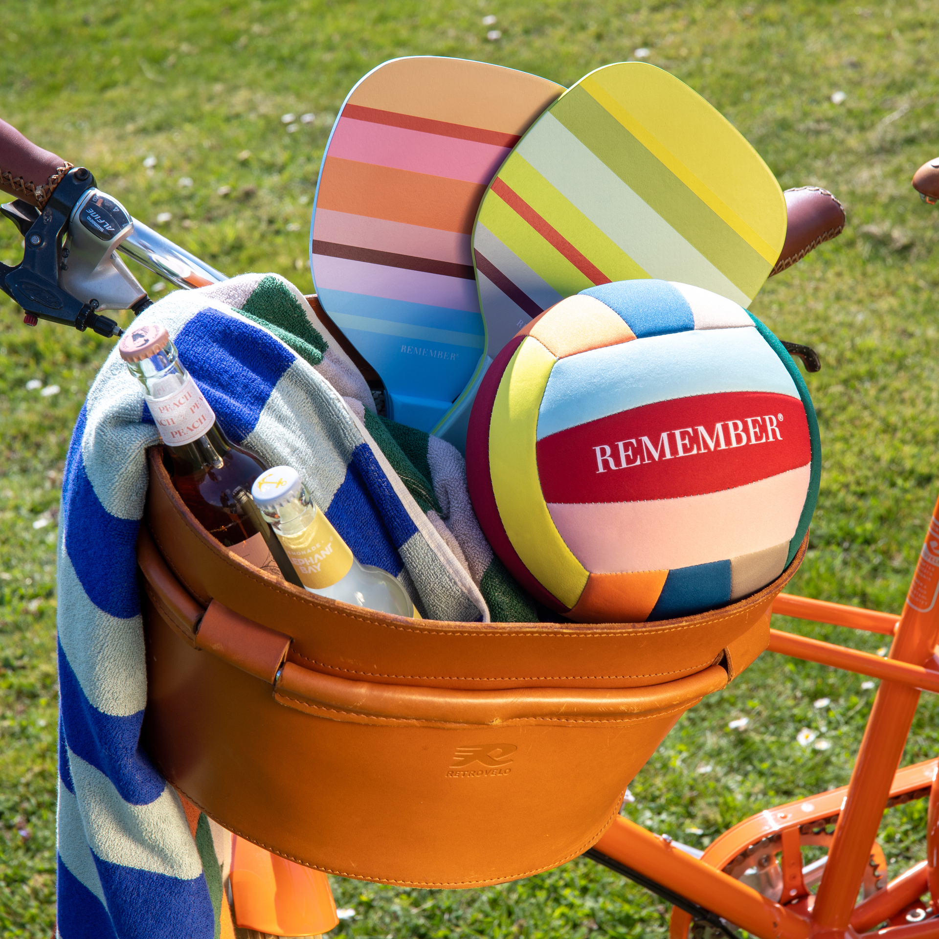 Fahrradkorb mit Beach Tennis-Set: Schläger, Ball, Getränke und Handtuch in bunten Streifen auf Rasen.