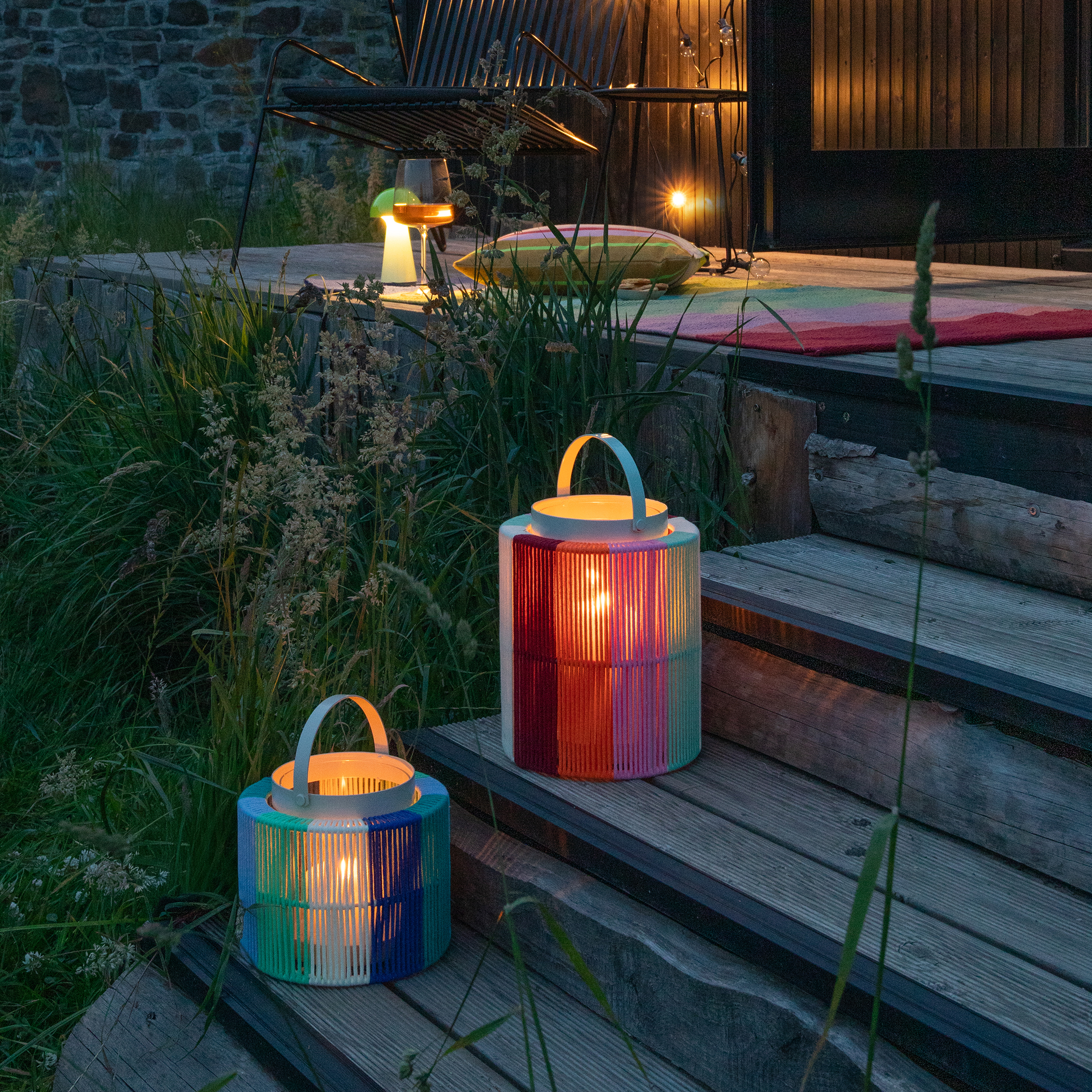 Zwei farbige Laternen auf Holzstufen. Im Hintergrund eine Terrasse mit Stuhl, Glas und einer Lampe. Der Boden ist mit einem bunten Teppich belegt. Umgeben von Gräsern.