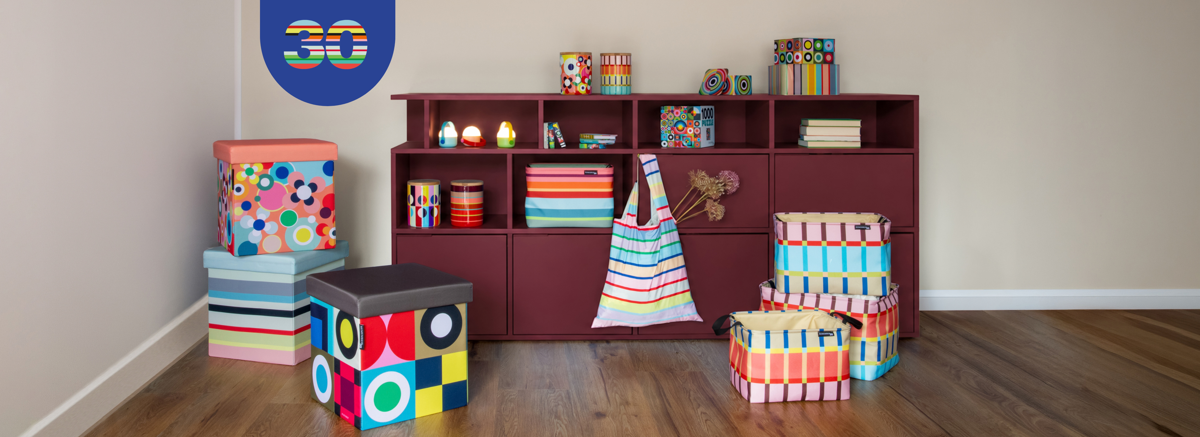 Pièce avec étagère rouge foncé et accessoires colorés : boîtes rayées et à motifs géométriques, livres, puzzle, tabourets et caisses aux motifs floraux, rayés et cubiques. Sac de courses suspendu. Logo 30 ans au mur.