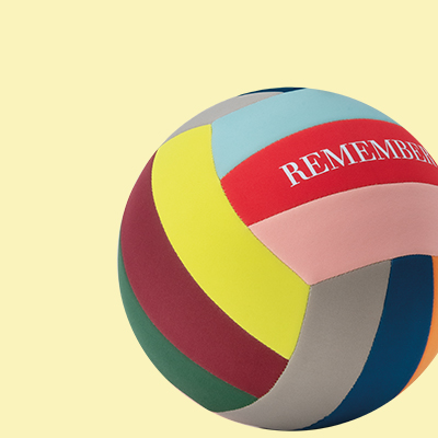 Ein bunter Volleyball mit dem Schriftzug "REMEMBER" auf einem roten Feld. Der Ball besteht aus Segmenten in Gelb, Rotbraun, Grün, Grau, Blau, Rosa und Türkis. Der Hintergrund ist gelb.