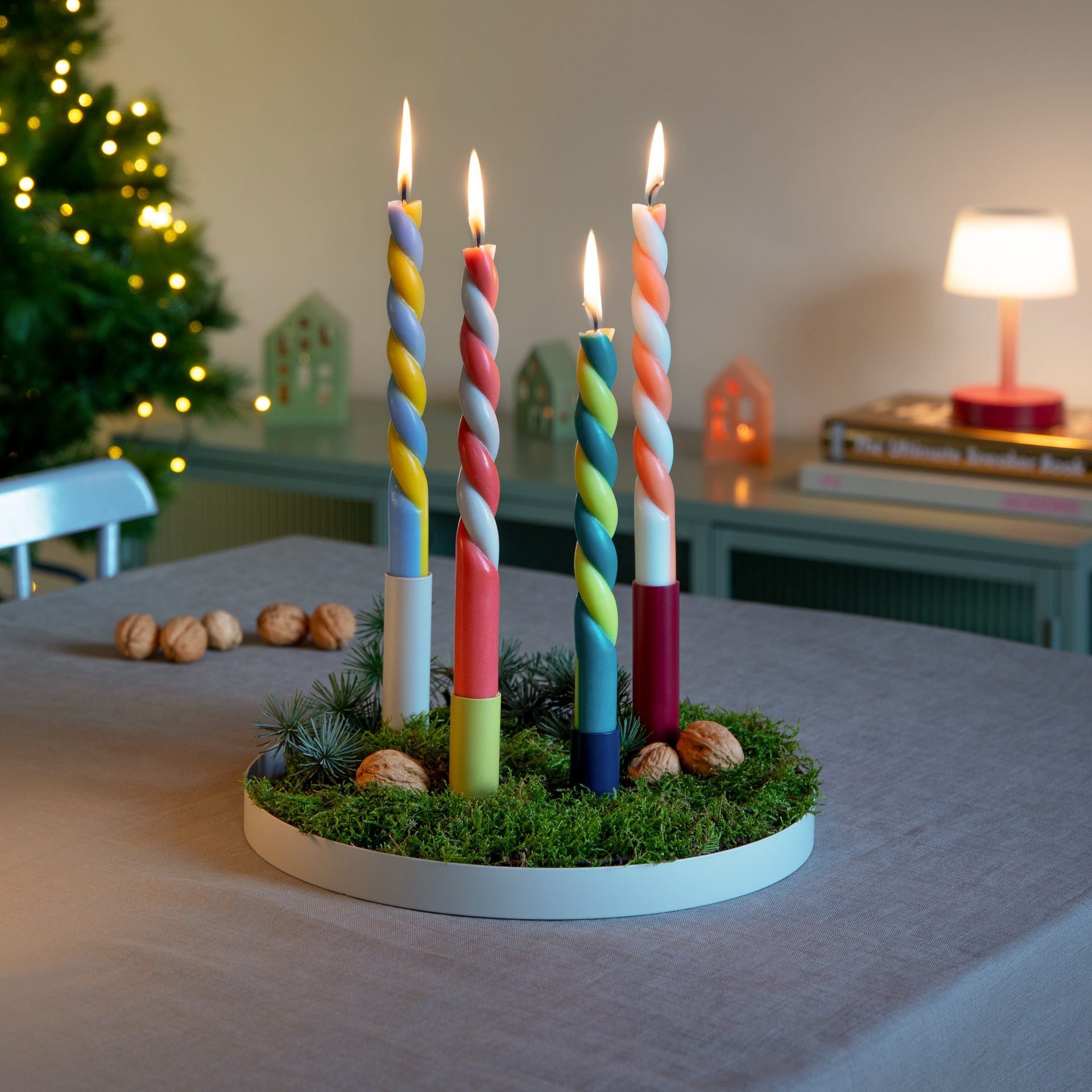 Magnetischer Kerzenhalter 'Luna' mit vier bunten, brennenden Kerzen auf einem runden Tablett mit Moos, Nüssen und Tannenzweigen, im Hintergrund ein Weihnachtsbaum.