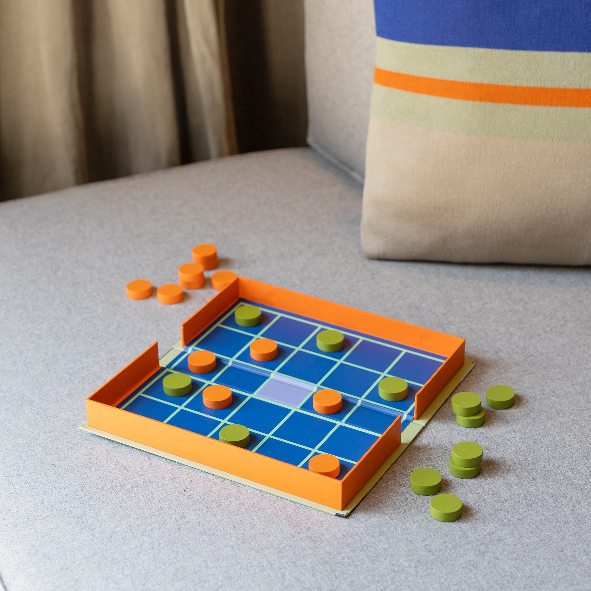 Verpackung des Seega Spiels. Abstraktes Design mit Kreisen, Linien und dem Spielnamen in orange und blau auf hellgrünem Hintergrund.