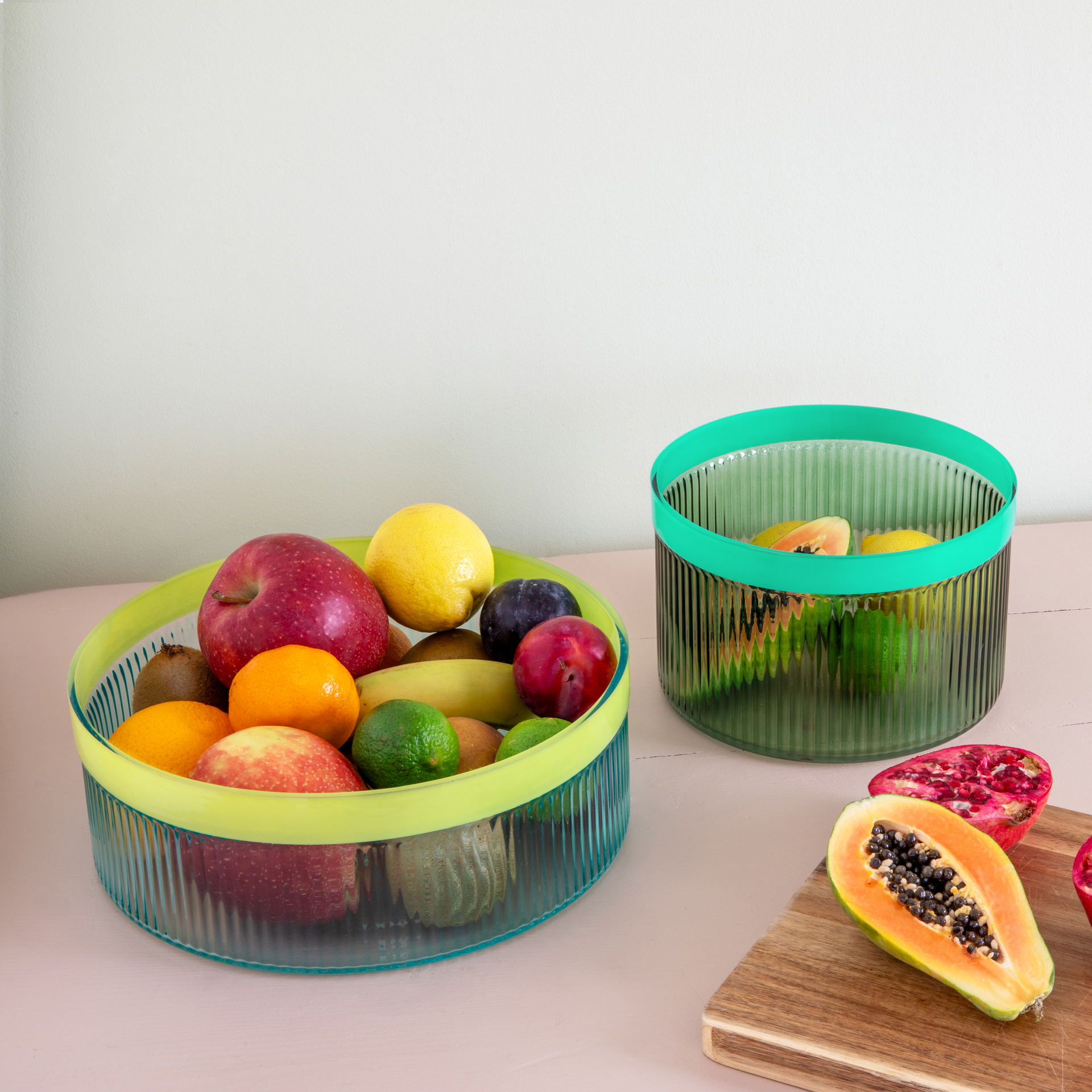 Zwei geriffelte Glasschalen, eine grün gefüllt mit Früchten wie Äpfel, Zitronen und Kiwis. Die andere Schale ist leer und daneben liegen Papaya und Granatapfel auf einem Holzbrett.