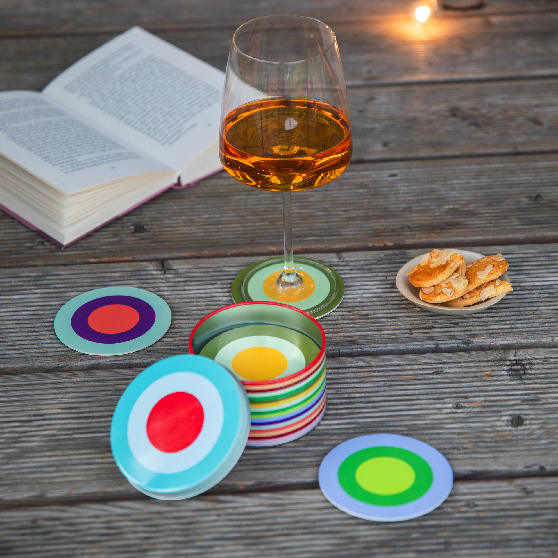Ein gedeckter Tisch mit Untersetzer-Set 'Ronda', einem Glas Wein, Keksen und einem Buch auf einer Holzoberfläche. Die Untersetzer sind bunt und kreisförmig.