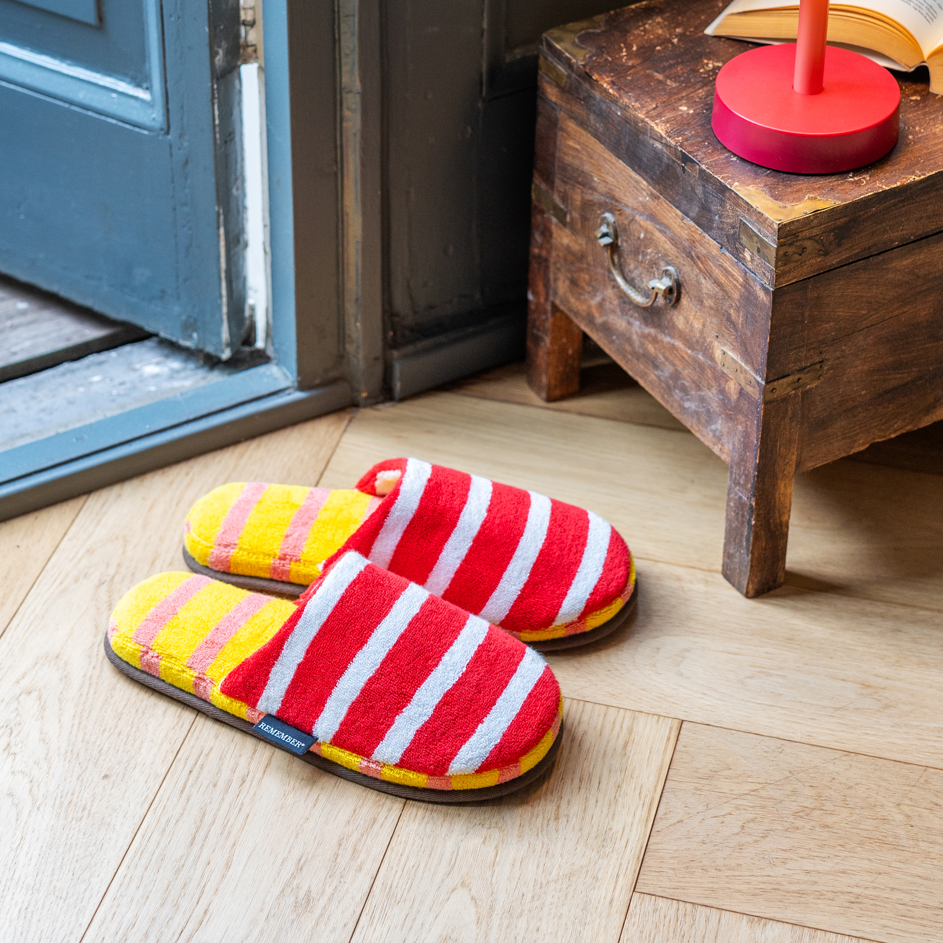 Zwei rote Mello Slipper mit weißen Streifen sind auf einem hellen Holzboden platziert, neben einer halboffenen blauen Tür und einer Holztruhe mit Lampe.