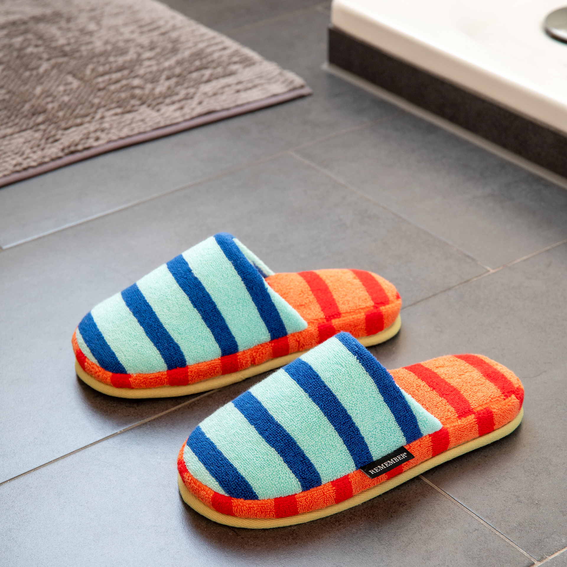 Zwei farbenfrohe, gestreifte Badeschuhe auf grauem Fliesenboden, ein Badteppich und ein Waschtisch im Hintergrund. Die Schuhe sind hellblau-dunkelblau und orange-rot gestreift.