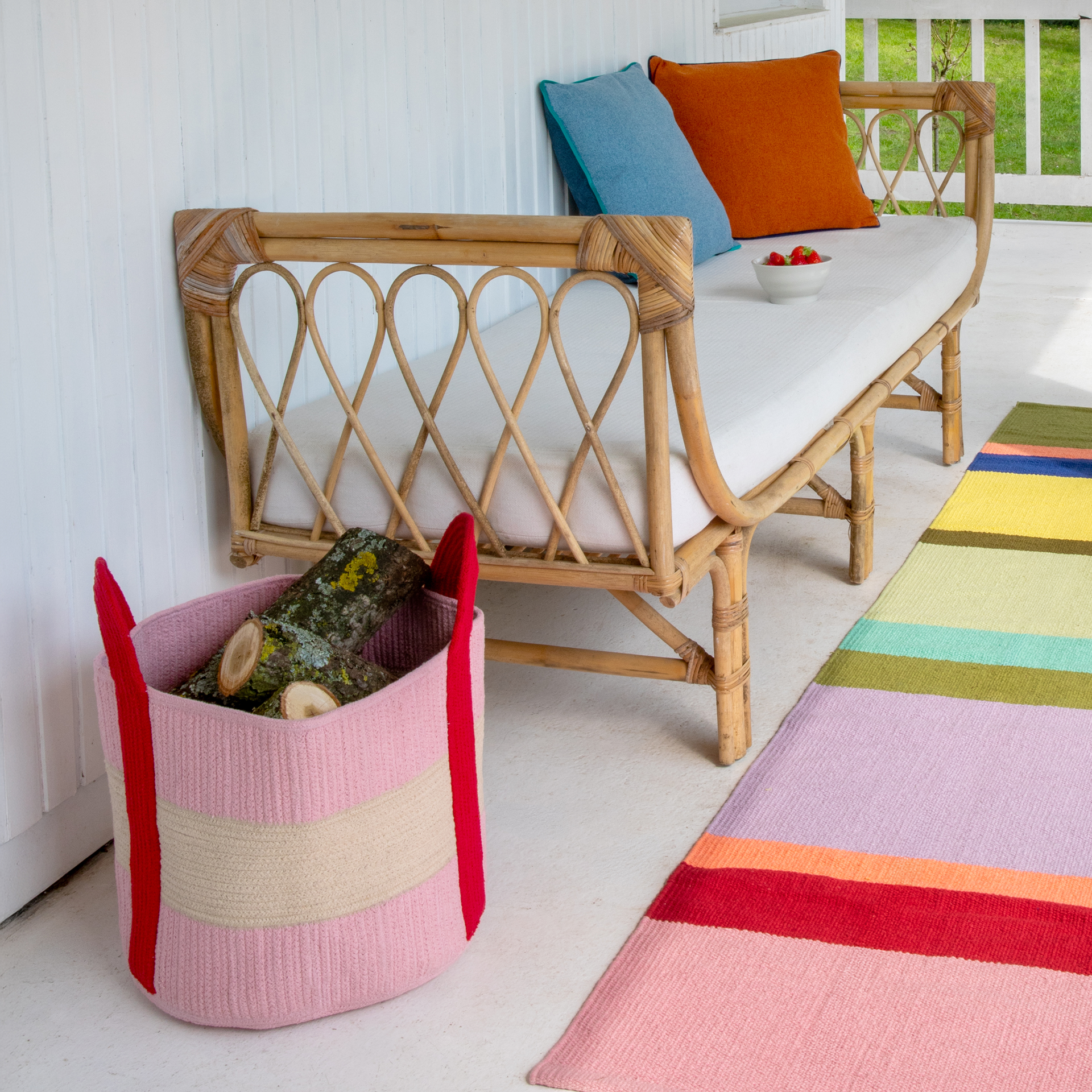 Gemütlicher Außenbereich mit Bambus-Tagesbett, weißer Auflage sowie blauem und orangem Kissen. Eine Schale Erdbeeren steht darauf. Daneben ein pinker Strickkorb mit Brennholz. Bunter Teppich auf hellem Boden.