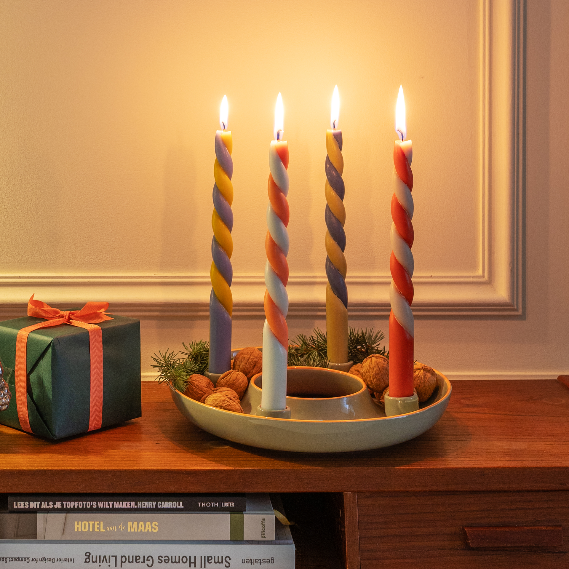 Adventskranzschale mit vier brennenden, farbig verdrehten Kerzen. Tannenzweige und Walnüsse liegen in der Schale. Geschenk daneben.