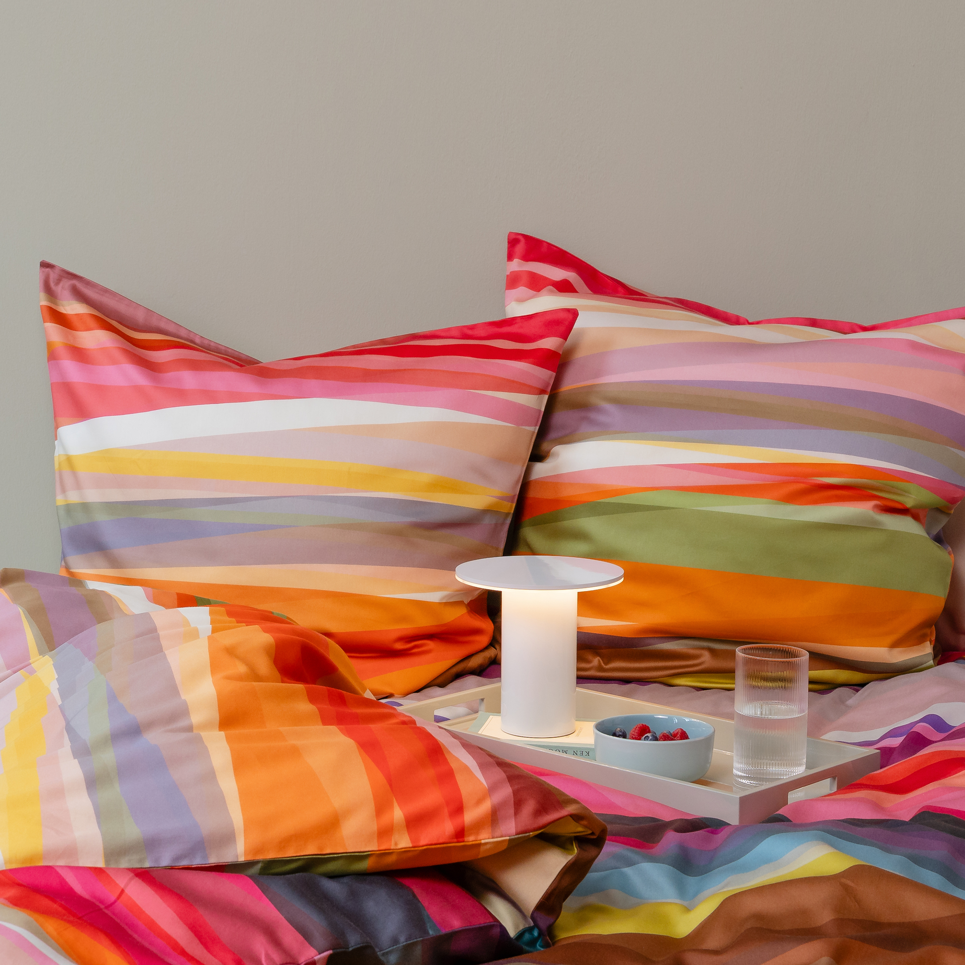 Detailansicht von 'Mateo' Bettwäsche (155x220 cm) mit bunten Streifen auf Kissen und Decke. Ein Nachttisch mit Lampe, Beeren und einem Glas Wasser steht auf dem Bett.