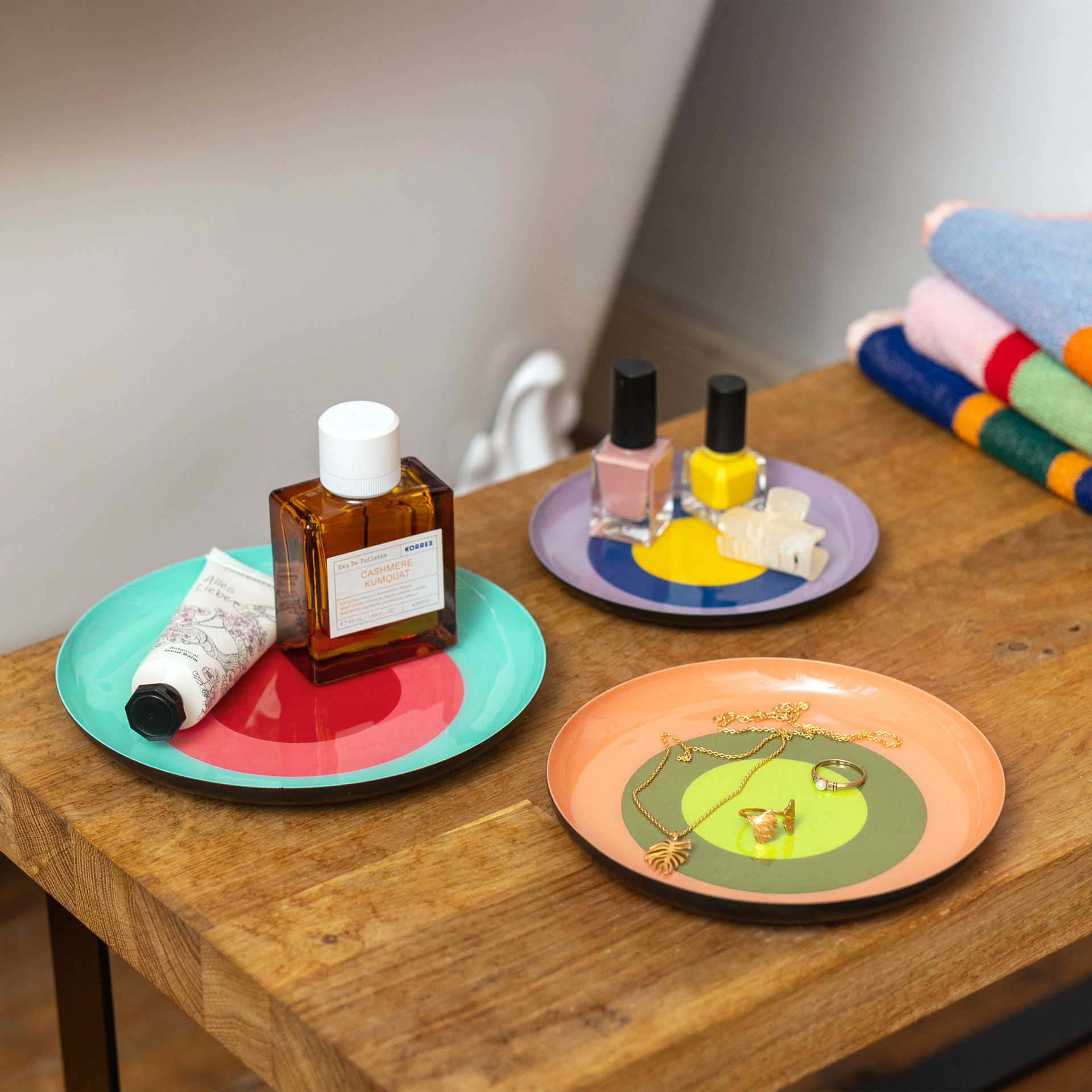 Drei farbige Ablageschälchen mit Kosmetik, Nagellack und Schmuck auf einem Holzuntergrund, gestreifte Handtücher im Hintergrund.
