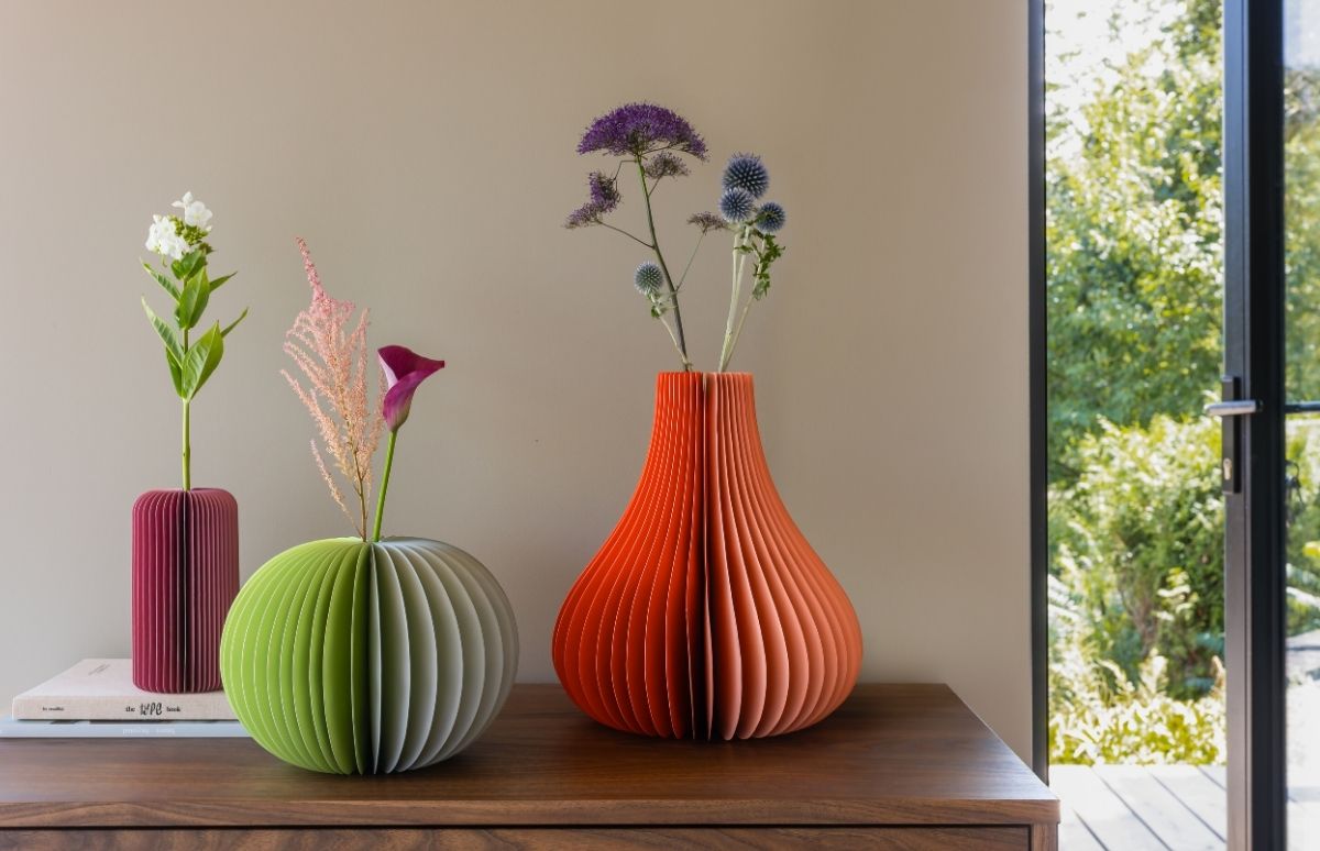 3 vases en papier plié sur une commode : cylindre magenta avec fleur blanche, boule verte avec fleurs roses, goutte orange avec chardons bleus. Fenêtre et verdure.