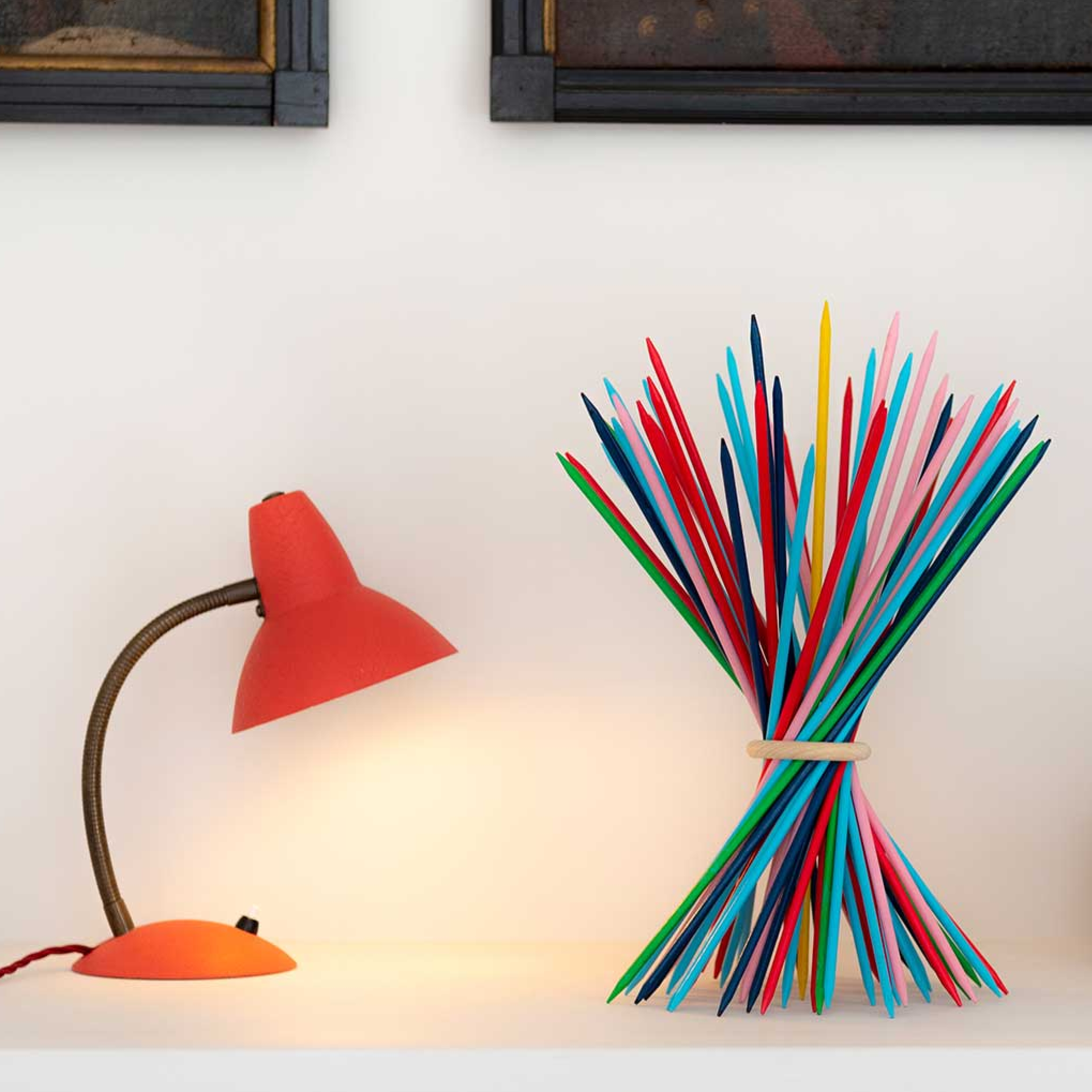 Dekorative, bunte Holzstäbe, zusammengebunden mit einem Holzring, neben einer roten Lampe auf einem Regal vor einer weißen Wand.