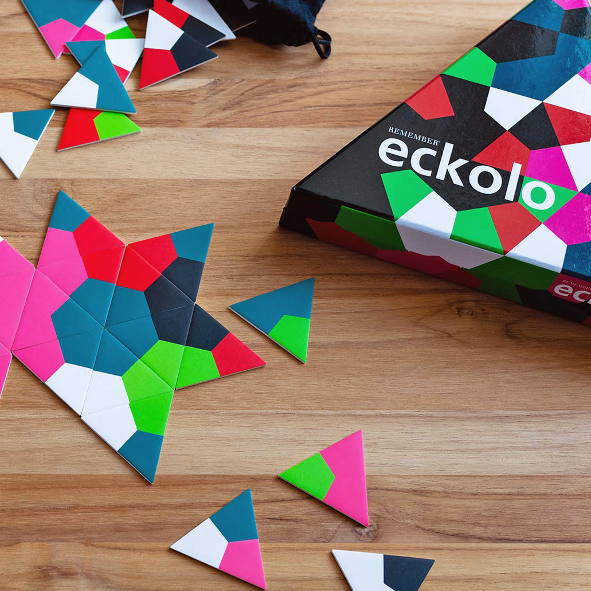 Eckolo Anlegespiel: Bunte Dreiecks-Karten auf Holzgrund, teils zu Stern gelegt. Eckige Spielbox rechts, mit dem 'eckolo' Schriftzug gut sichtbar.
