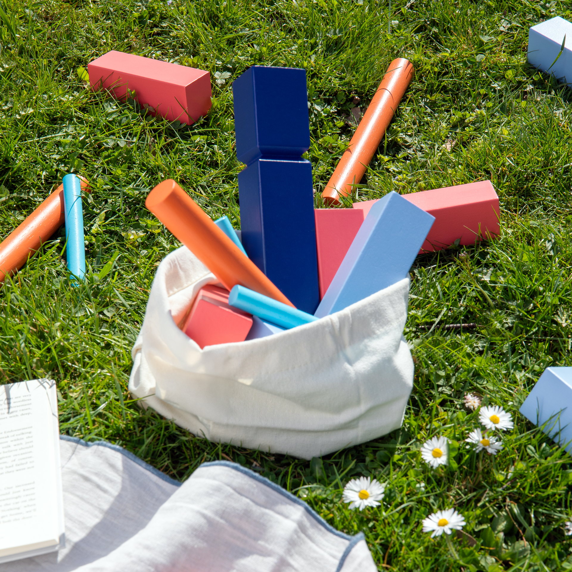 Draufsicht von Kubb-Spielsteinen aus Holz in verschiedenen Farben, teilweise im Stoffbeutel auf einer Wiese. Ein Buch und Gänseblümchen sind ebenfalls sichtbar.