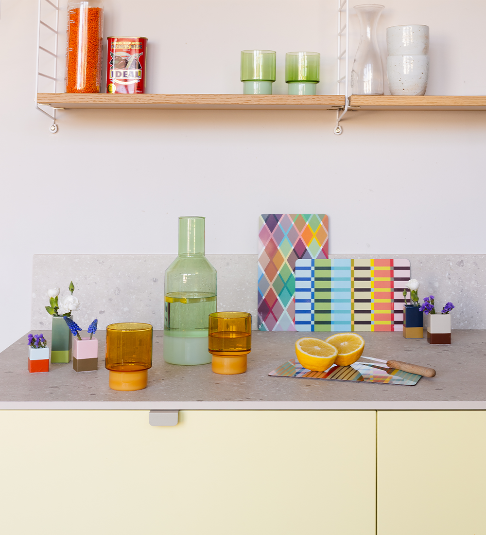 Helle Küche mit Glaswaren, Deko und Zitrone auf einer Arbeitsfläche. Im Hintergrund ein Regal mit Lebensmitteln und weiteren Gläsern. Farblich abgestimmtes Küchenzubehör.