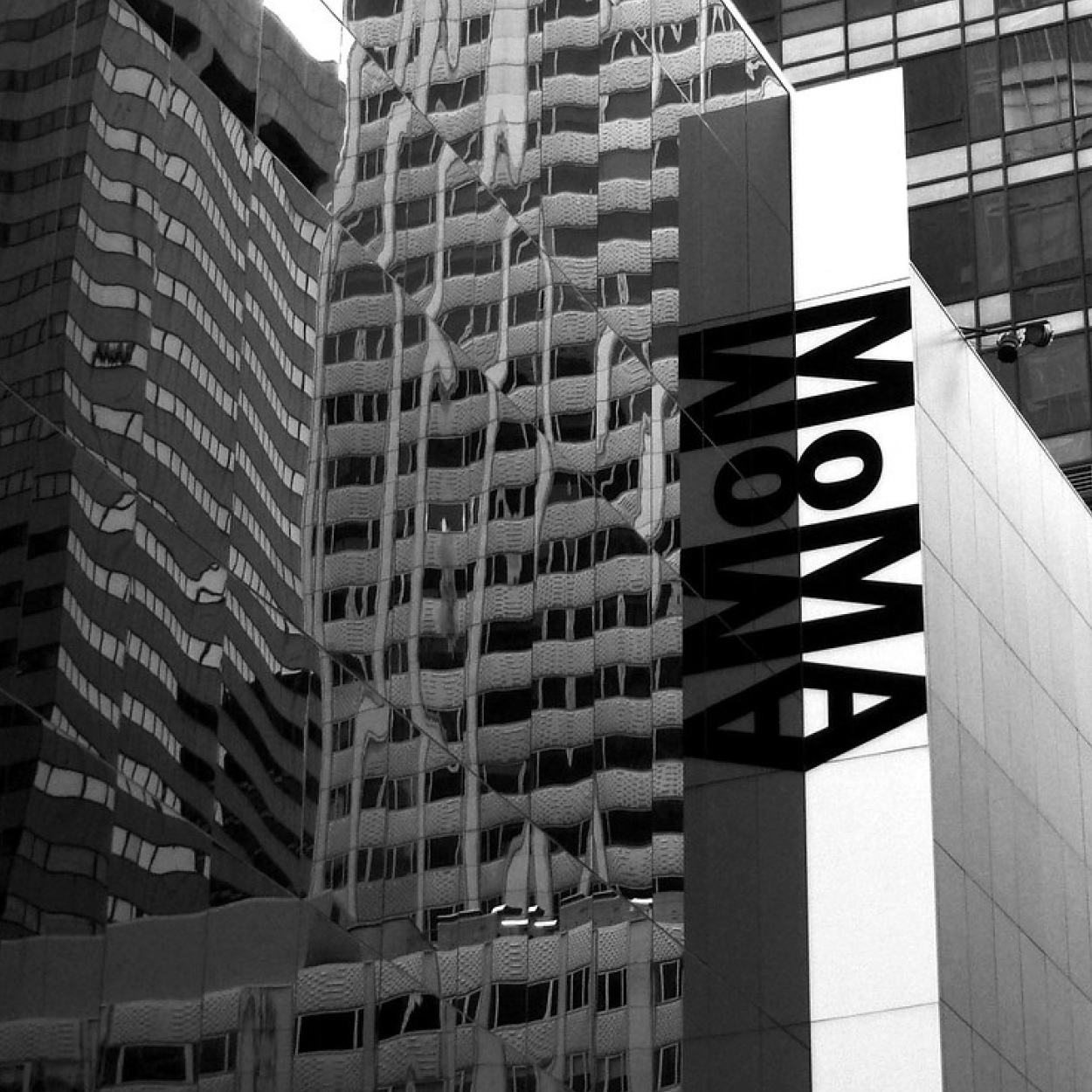 Schwarzweiß-Aufnahme des MoMA-Gebäudes. Spiegelfassade reflektiert umliegende Gebäude mit wellenförmigen Fassaden. Das MoMA-Logo ist senkrecht auf der Gebäudefassade angebracht.