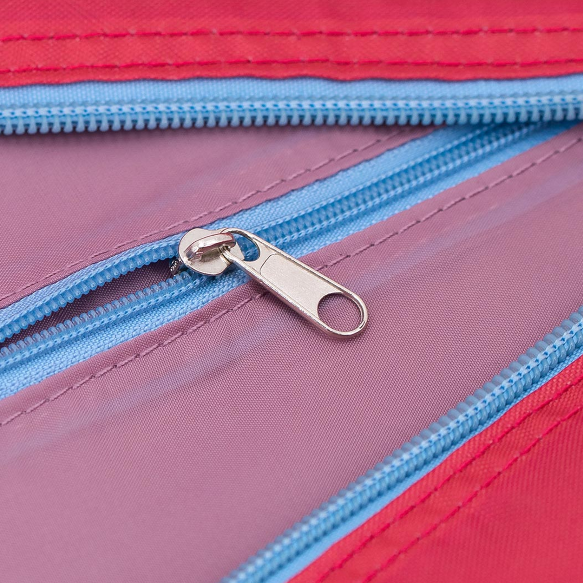 Detailaufnahme der 'Ida' Reisetasche mit Reißverschluss. Der Zipper ist aus Metall und liegt auf rosafarbenem Stoff. Umgeben ist der Stoff mit einem hellblauen Reißverschluss und rotem Stoff.