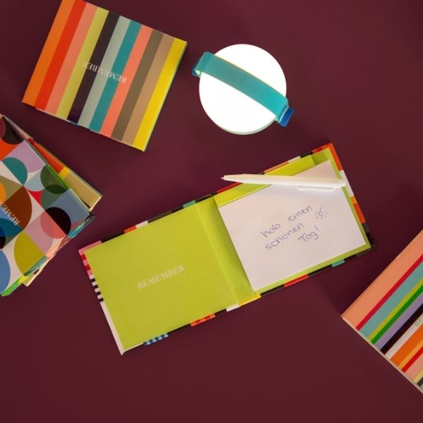 Des carnets colorés sur fond rouge foncé. Un livre ouvert montre une page vert tilleul «REMEMBER » et un petit mot « Passe une bonne journée ! » avec un soleil. Un stylo blanc y est planté. Lampe ronde avec ruban bleu.