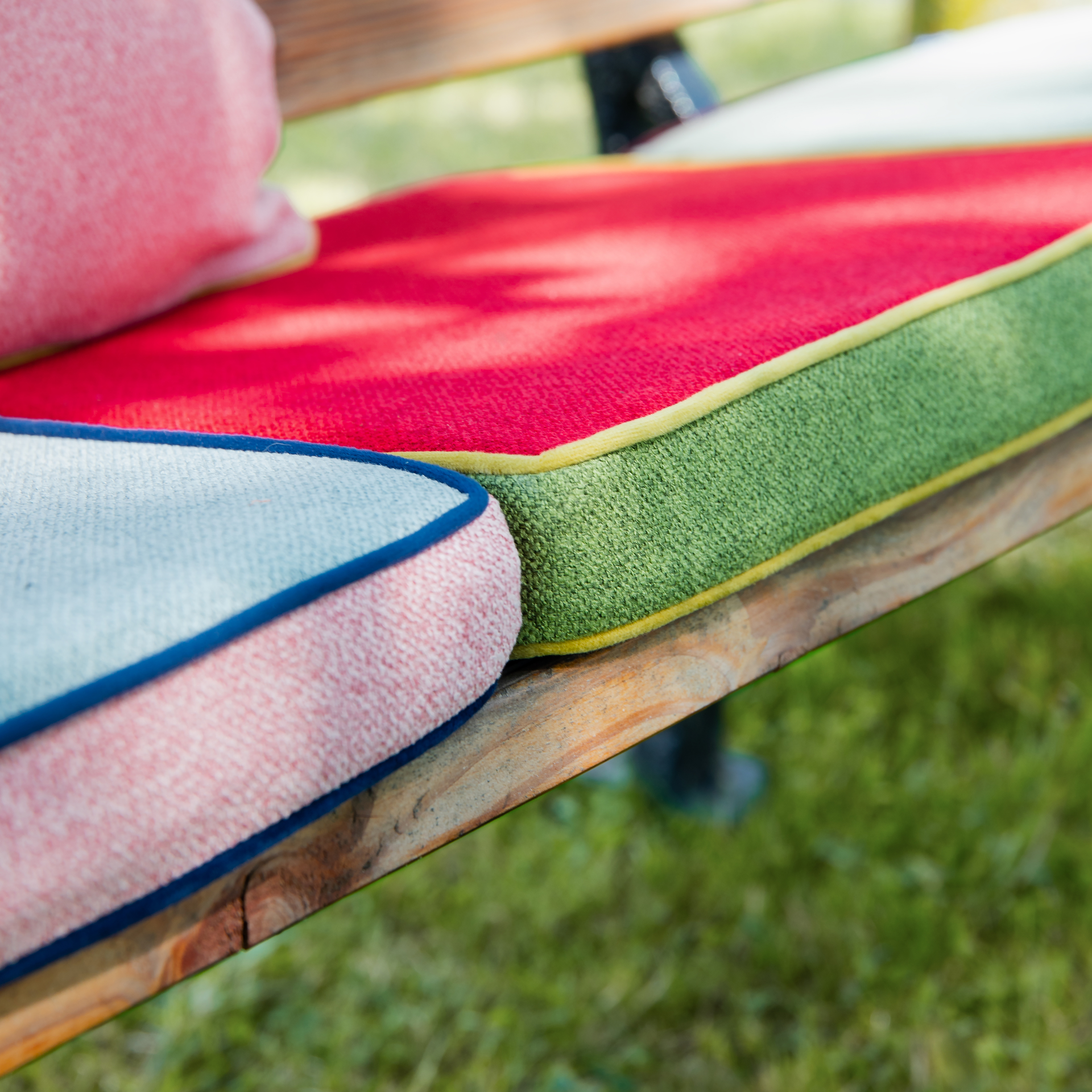 Detailaufnahme eines mehrfarbigen Sitzkissens mit Paspelierung auf einer Holzbank. Die Farben umfassen Cranberry-Rot, Grün, Rosa und Blau.