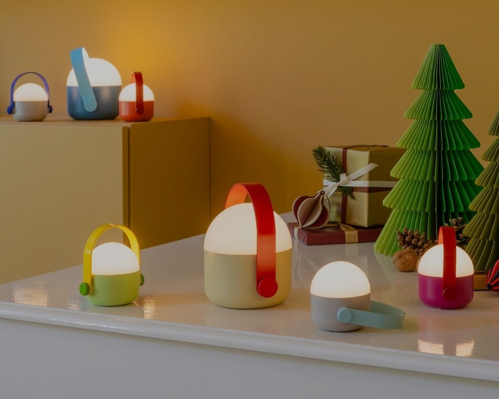 Sieben stilvolle, tragbare LED-Lampen mit farbigen Griffen in verschiedenen Größen und Farben wie Hellgrün, Gelb, Rot, Blau, Grau und Magenta. Warmes Licht auf weißem Sideboard und Regal; im Hintergrund grüne Papierbäume, Geschenke und Zapfen.