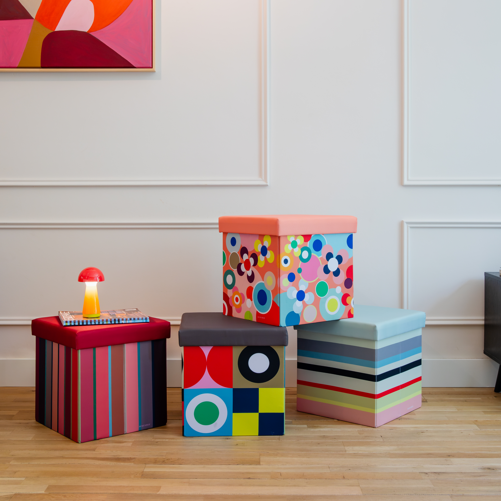 Quatre coffres-sièges colorés sont posés sur un plancher en bois. Les coffres « REMEMBER » arborent des rayures ainsi que des motifs floraux et géométriques. Ils servent de sièges et offrent un espace de rangement pratique.