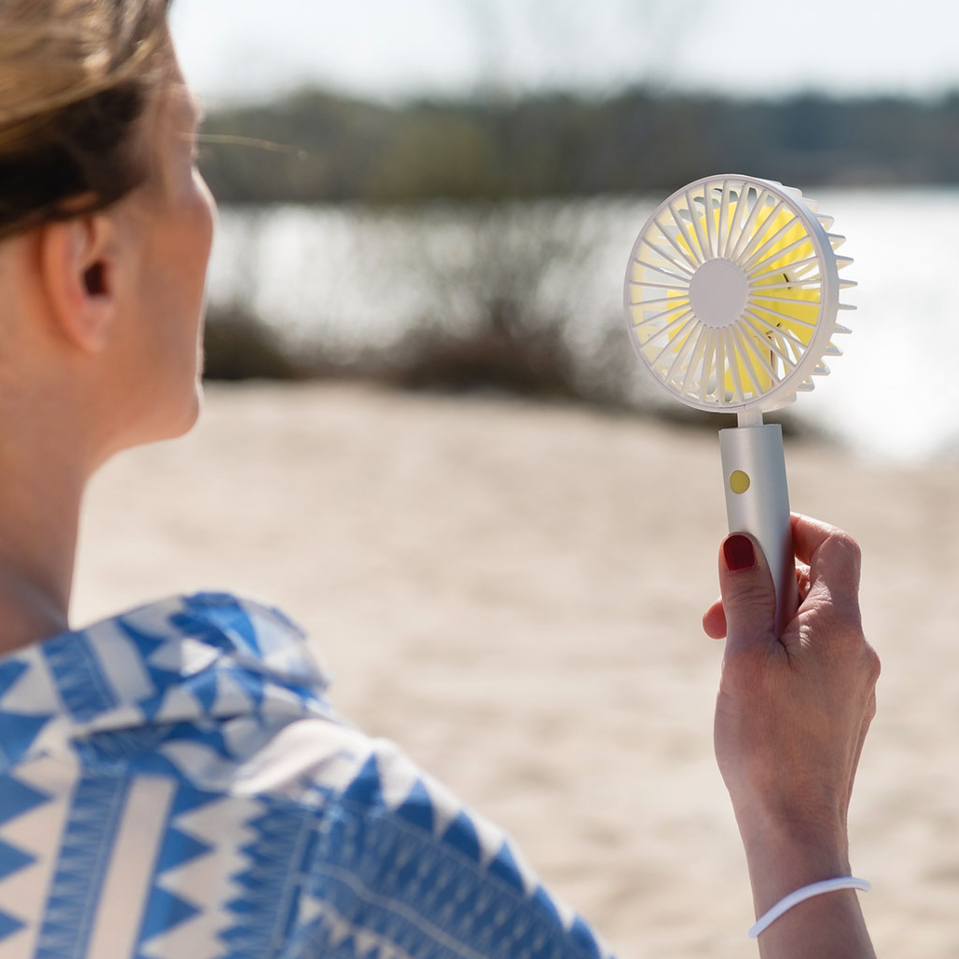Frau hält silbernen Akku-Ventilator 'Flow' mit gelben Rotorblättern vor Strandkulisse in der Hand.