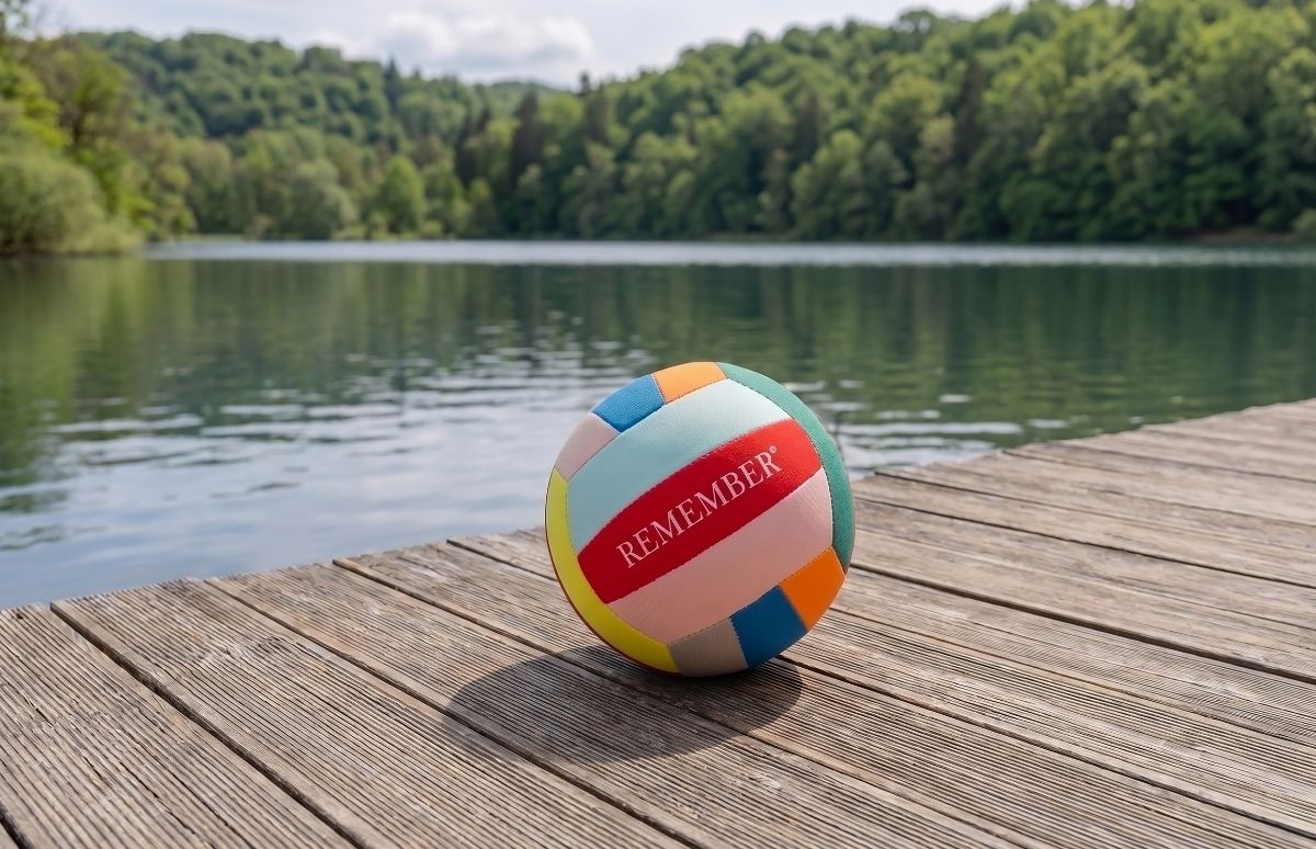 Una palla da pallavolo colorata con la scritta rossa "REMEMBER" giace su un pontile di legno al sole. La palla ha toni pastello: rosso, blu, giallo e verde. Sullo sfondo, un lago e una fitta foresta.