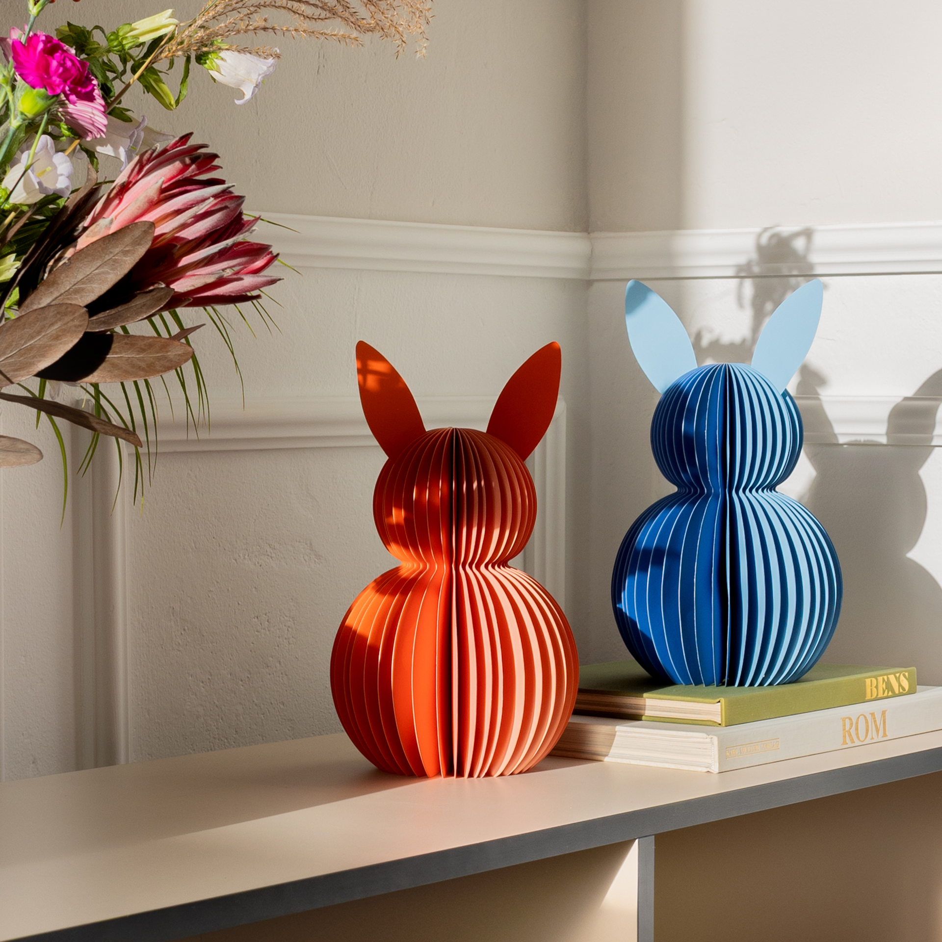 Zwei Papierhasen: links blau, rechts orange. Jeder Hase besteht aus gefächertem Papier und hat Ohren.