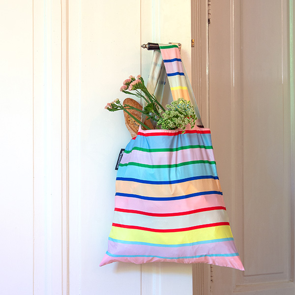 Un sac à provisions multicolore à rayures est accroché à une poignée de porte. Une baguette croustillante et des fleurs fraîches dans des tons de rose et de vert clair dépassent du sac. Rayures pastel dans les tons de rose, bleu, jaune et vert.