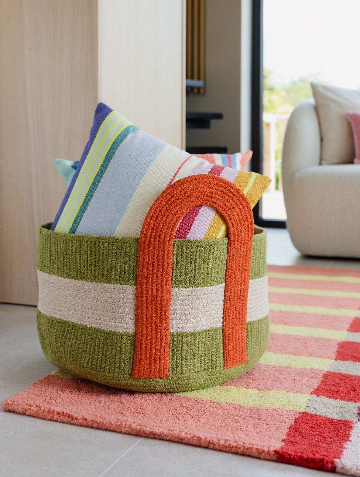 Panier rond tressé vert, orné d'une bande beige et d'une poignée en forme de U orange, posé sur un tapis aux couleurs vives. Dans le panier, plusieurs coussins rayés aux couleurs pastel claires. Décor intérieur lumineux.
