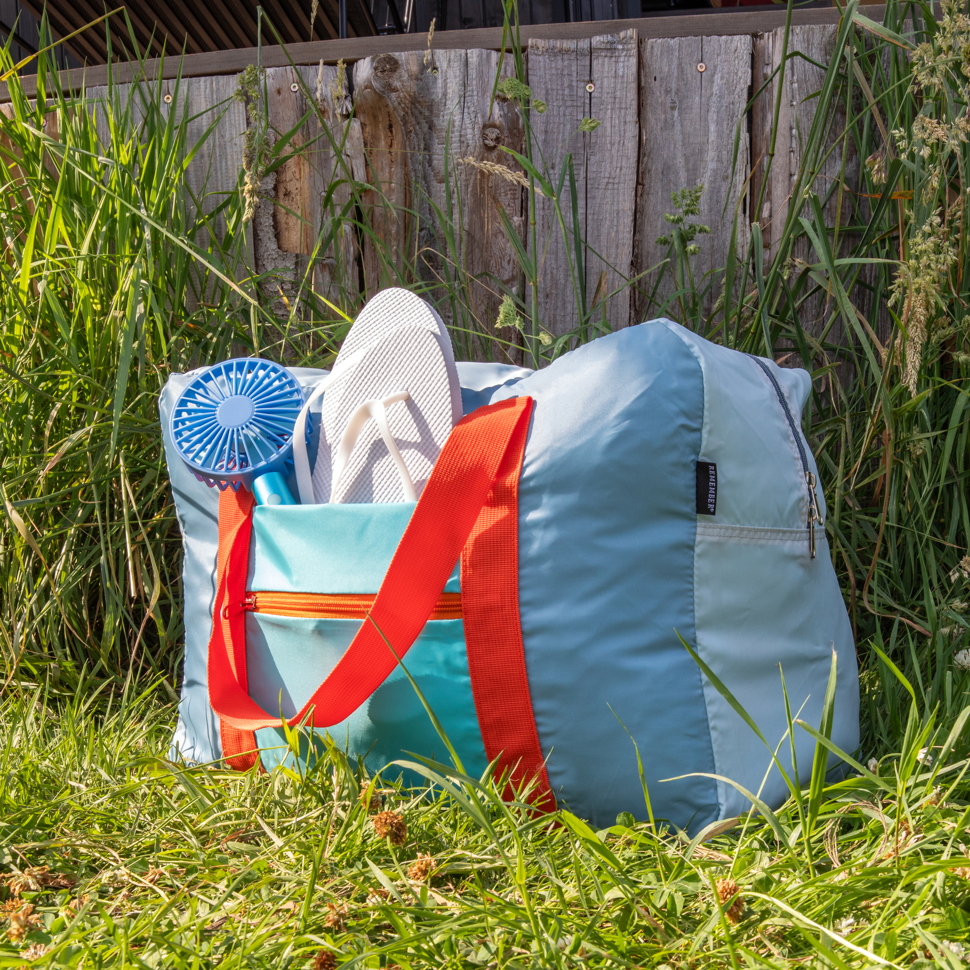 Hellblaue Reisetasche 'Tom' mit orangen Trägern im Gras. Darin: Weisse Flip-Flops und ein Mini-Ventilator. Im Hintergrund ein Holzzaun.