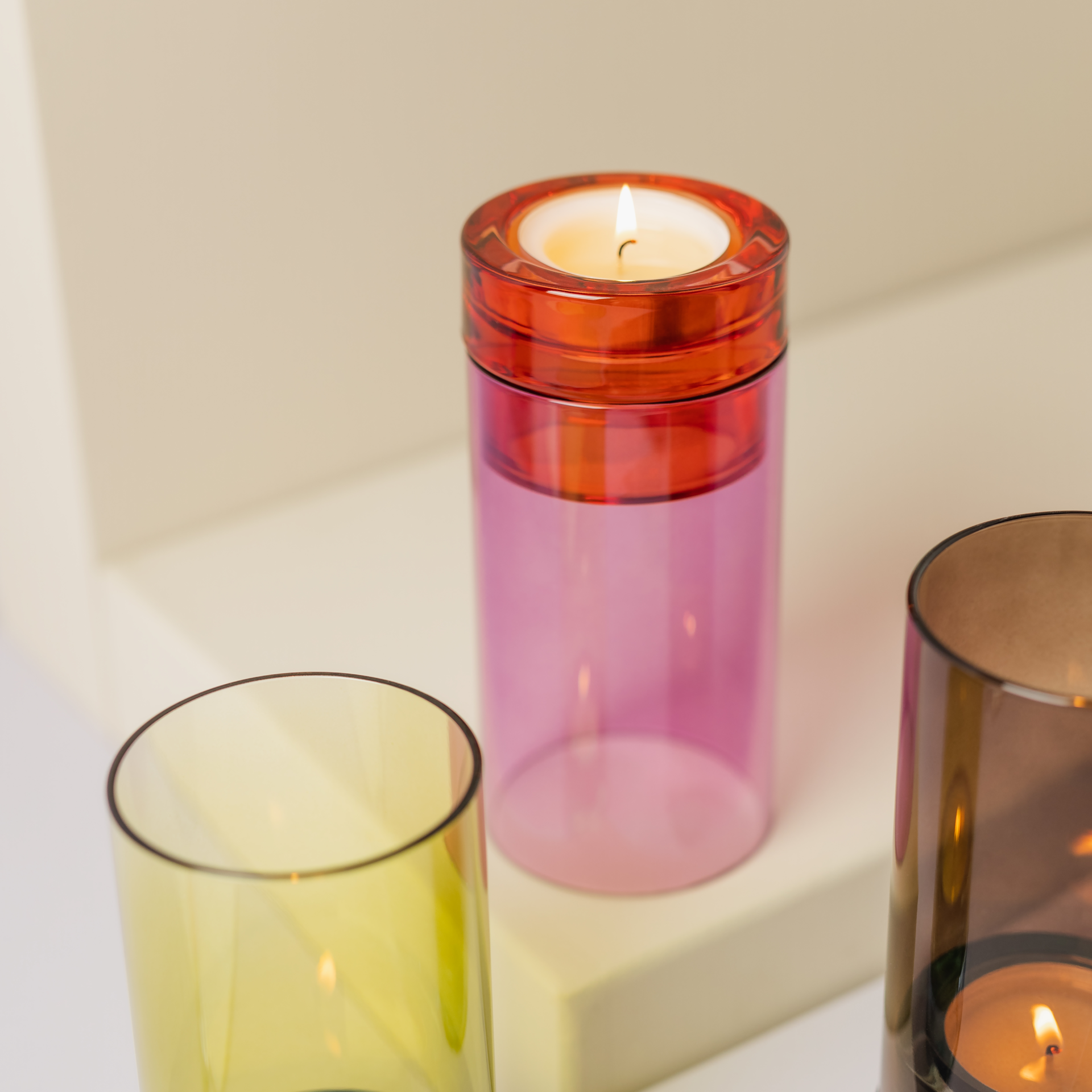 Pinkes Glaswindlicht mit Teelicht, zylindrischer Form. Sockel in Orange, darauf ein Einsatz für das Teelicht. Das Teelicht steht im pinkfarbenen Glaszylinder.