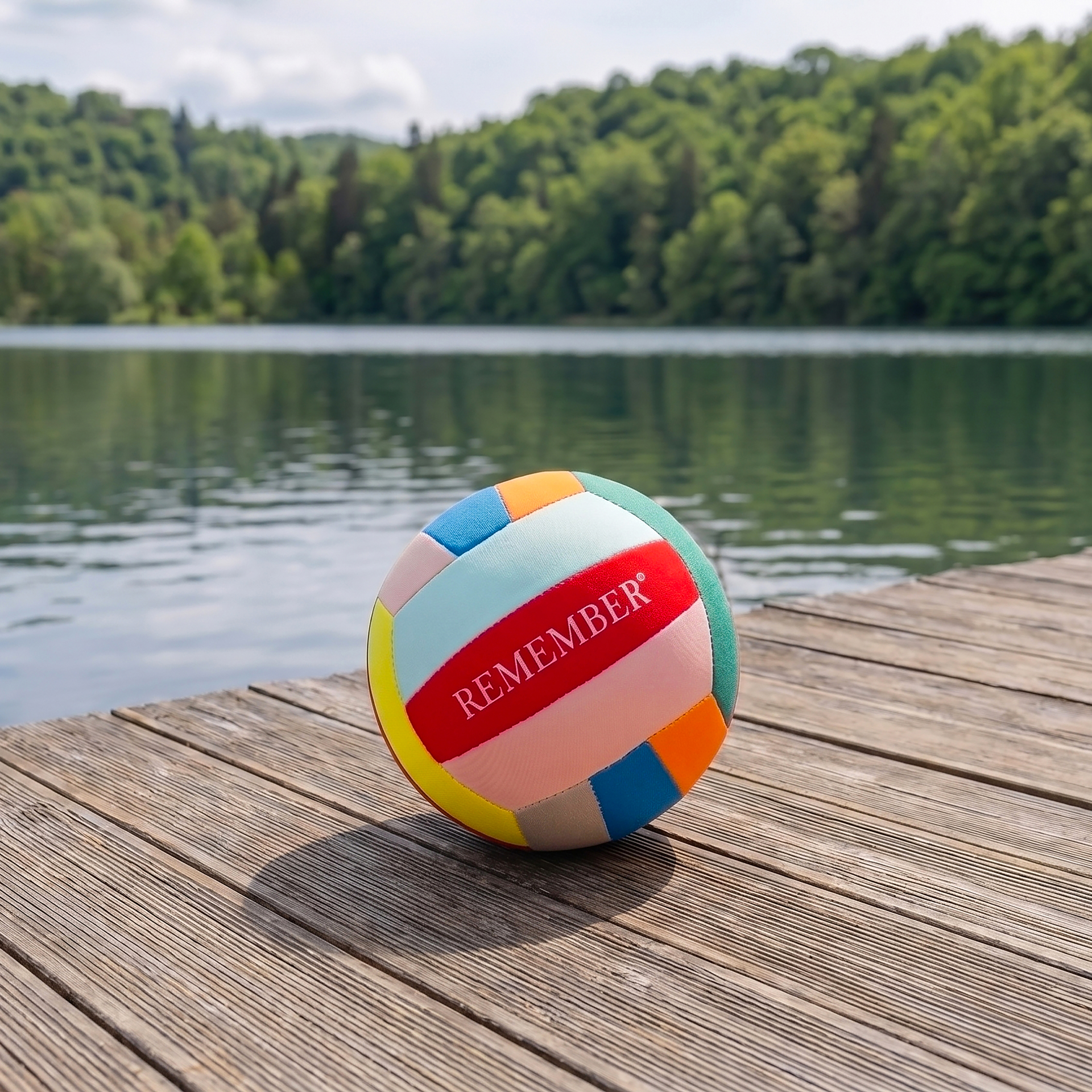 Ein bunter Ball, ähnlich einem Strand- oder Volleyball, mit dem Wort REMEMBER in Rot auf hellrosa Feld, liegt auf einem sonnenbeschienenen Holzsteg. Dahinter ein ruhiger See, umgeben von grünen Wäldern unter leicht bewölktem Himmel.