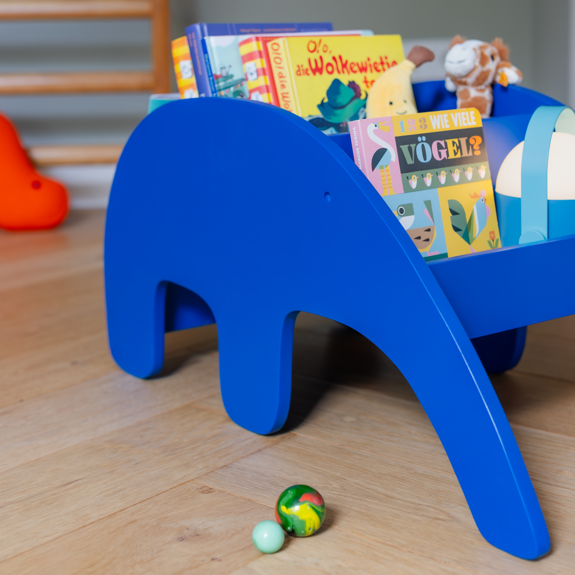 Ein blaues, elefantenförmiges Bücherregal steht auf einem hellen Holzboden. Das Regal ist mit Büchern und Stofftieren gefüllt. Im Vordergrund liegen Murmeln.