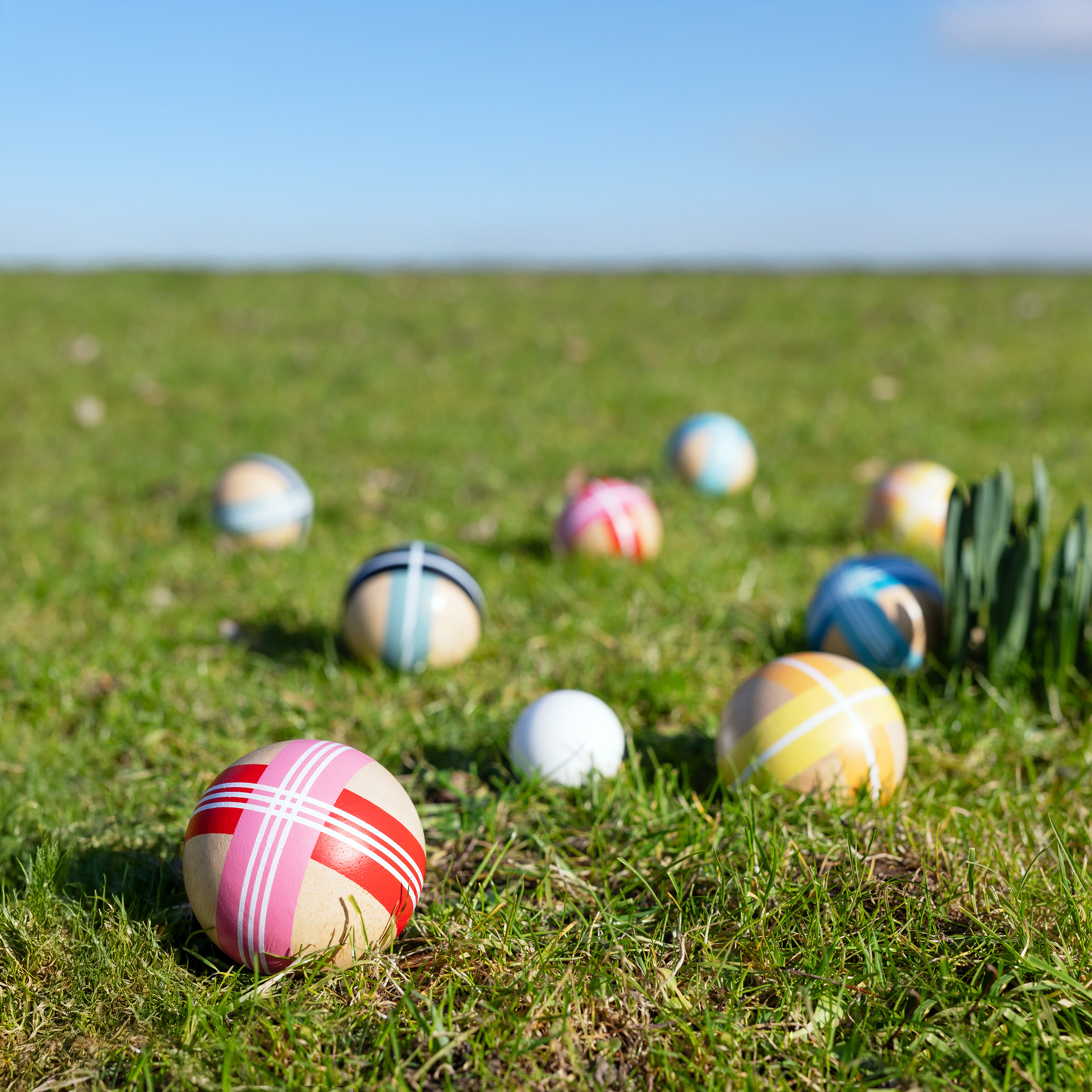 Grüne Wiese mit bunten Boccia-Kugeln. Holz-Kugeln mit Streifenmuster liegen verstreut im Gras, blauer Himmel im Hintergrund.