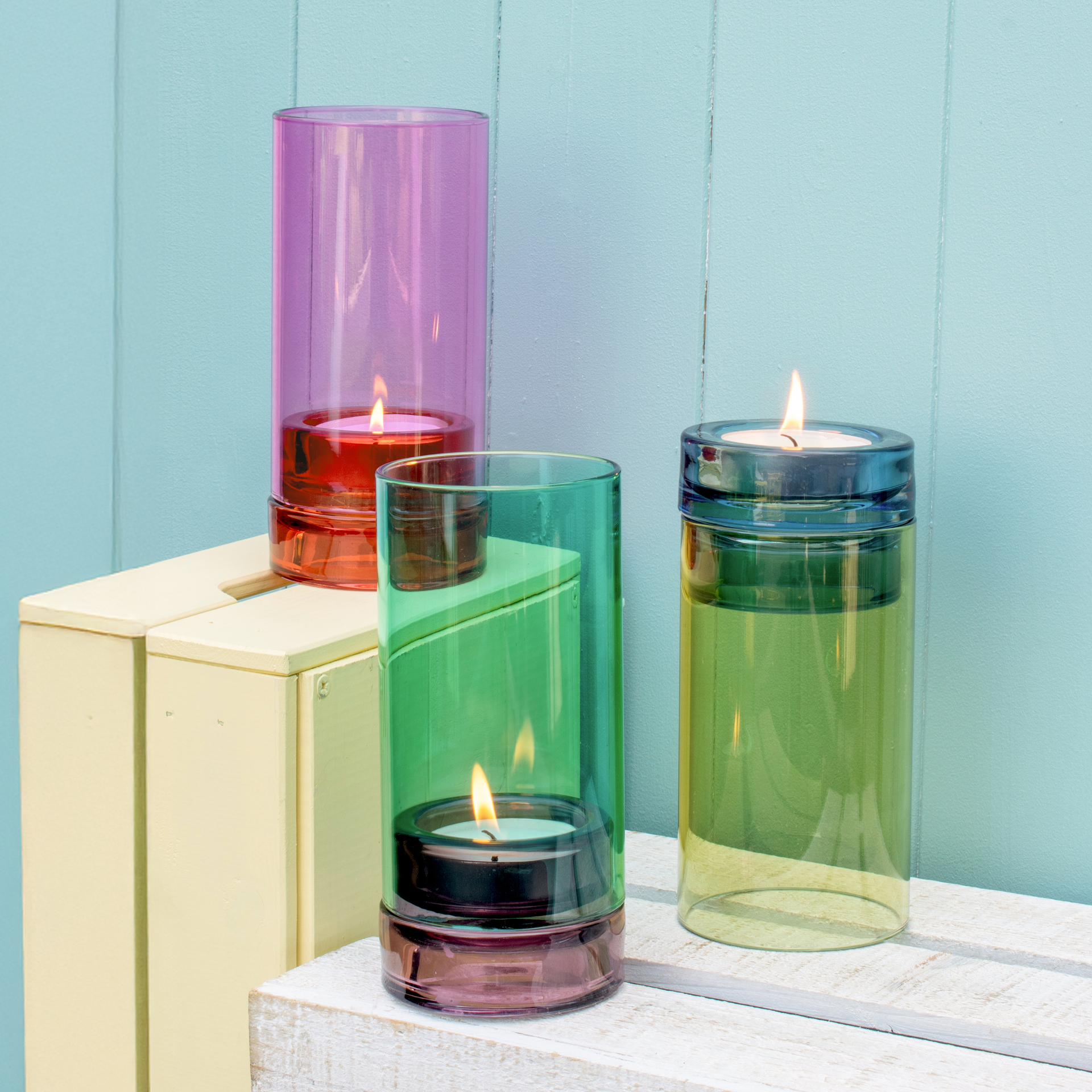 Drei Glas-Windlichter in Aqua, Grün und Pink mit brennenden Teelichtern auf Holzkisten vor türkisem Hintergrund.