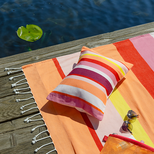 Ein farbenfrohes, gestreiftes Badetuch mit passendem Kissen, Sonnenbrille und Netztasche liegt auf einem Holzanleger am Wasser. Leuchtende Farben wie Orange, Pink und Rot kontrastieren mit dem dunklen Wasser, in dem ein Seerosenblatt schwimmt.