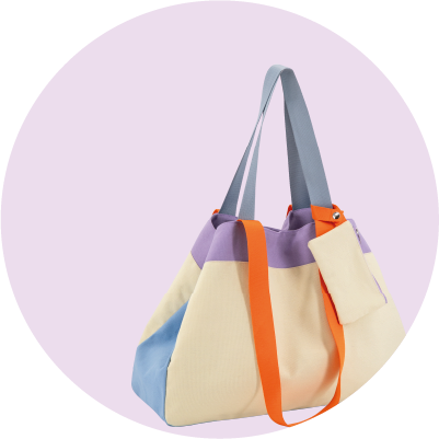 Bunte Stofftasche in hellen Farben auf lila Hintergrund. Beigefarbener Stoff mit Lavendelstreifen oben und hellblauen Seitenteilen unten. Graublaue Griffe, orange Riemen und kleine beigefarbene Zusatztasche. Modern.