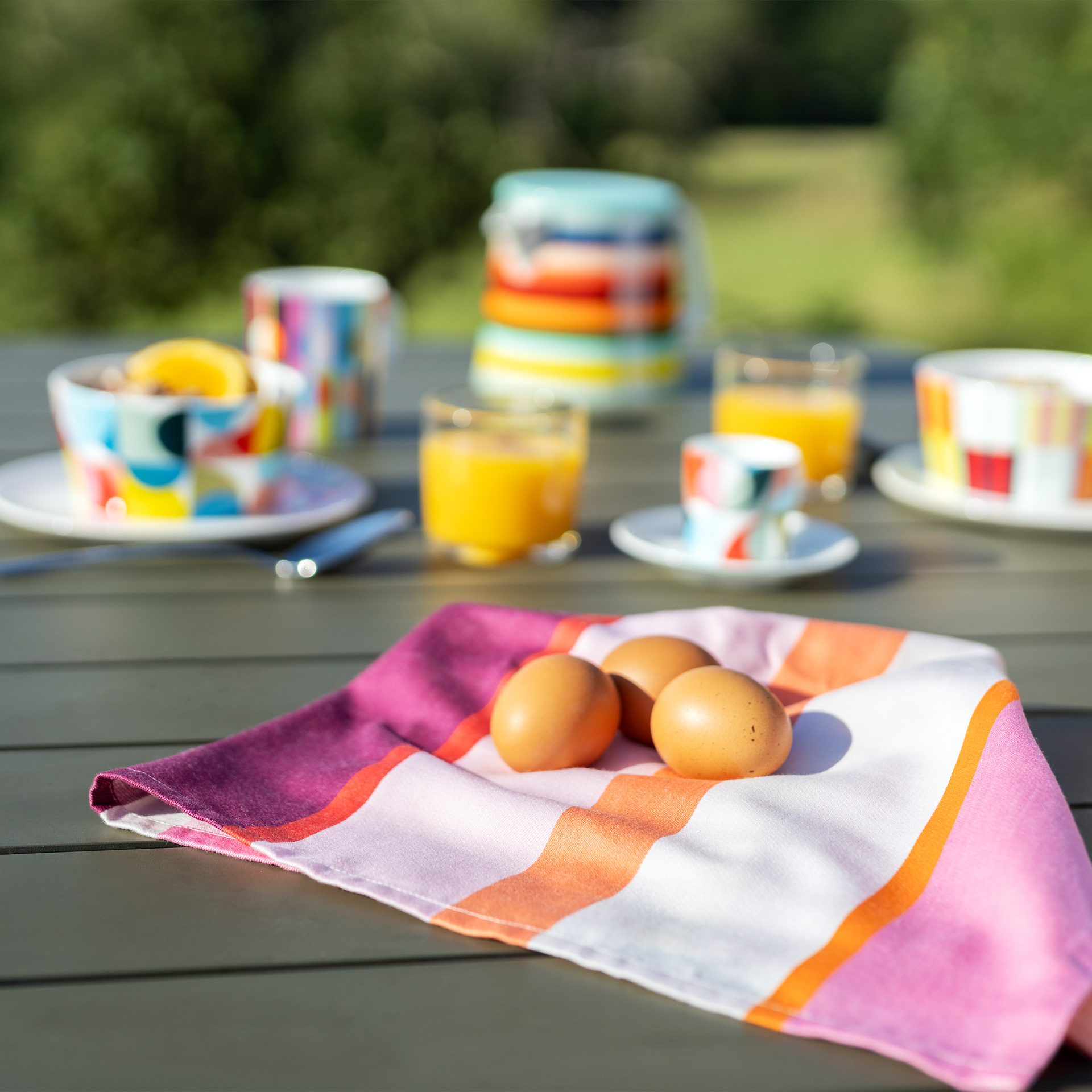 Ein farbenfrohes Trockentuch mit breiten Streifen in Pink, Orange, Lila und Weiß liegt auf einem dunklen Holztisch. Drei braune Eier ruhen darauf. Im unscharfen Hintergrund buntes Frühstücksgeschirr und Orangensaft im Freien.