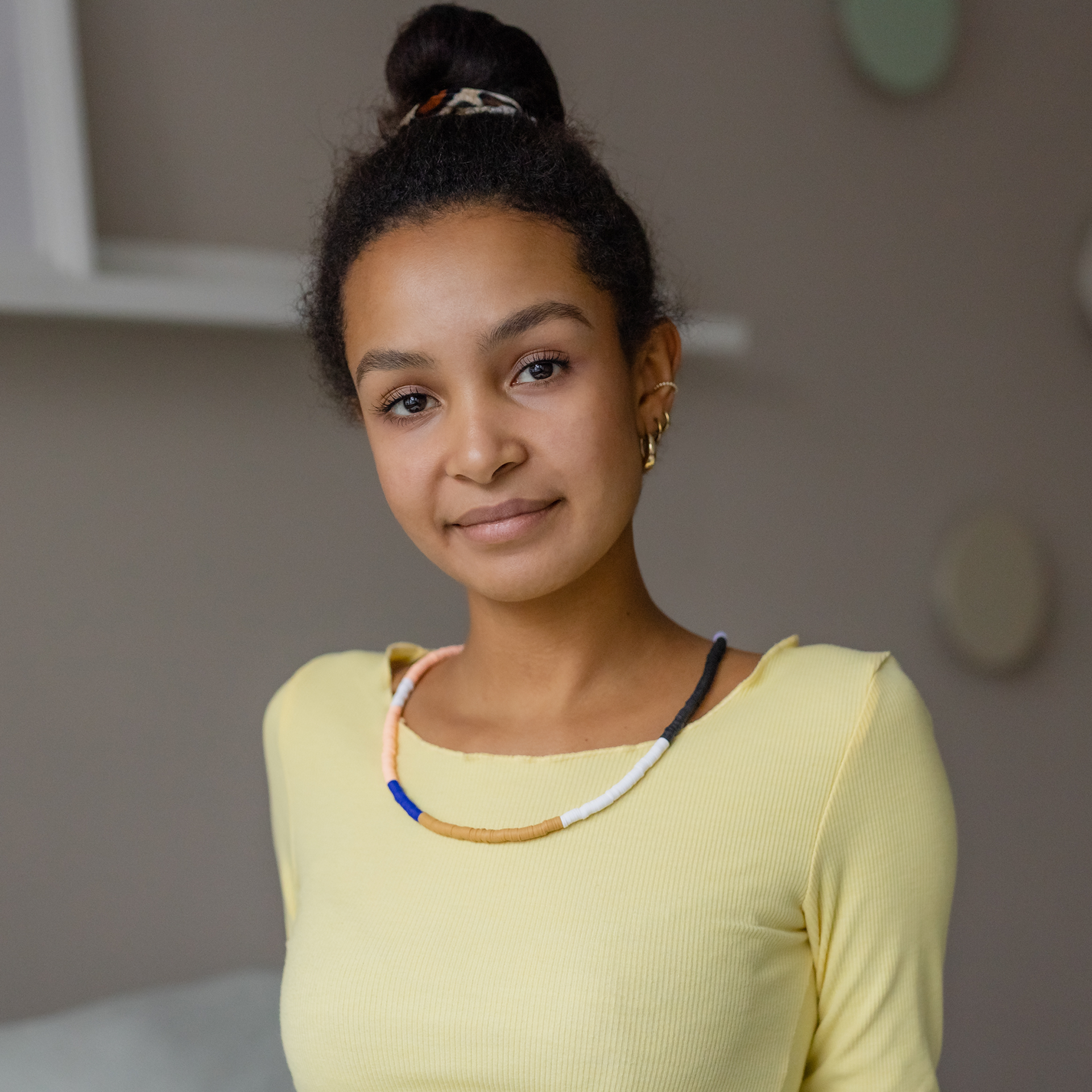 Junge Frau mit dunklem Teint und zum Dutt gebundenen Haaren blickt freundlich direkt in die Kamera. Sie trägt eine pastellfarbene Perlenkette in Rosa, Blau, Beige, Weiß und Dunkelgrau über hellgelber, gerippter Bluse. Dezente goldene Ohrringe.