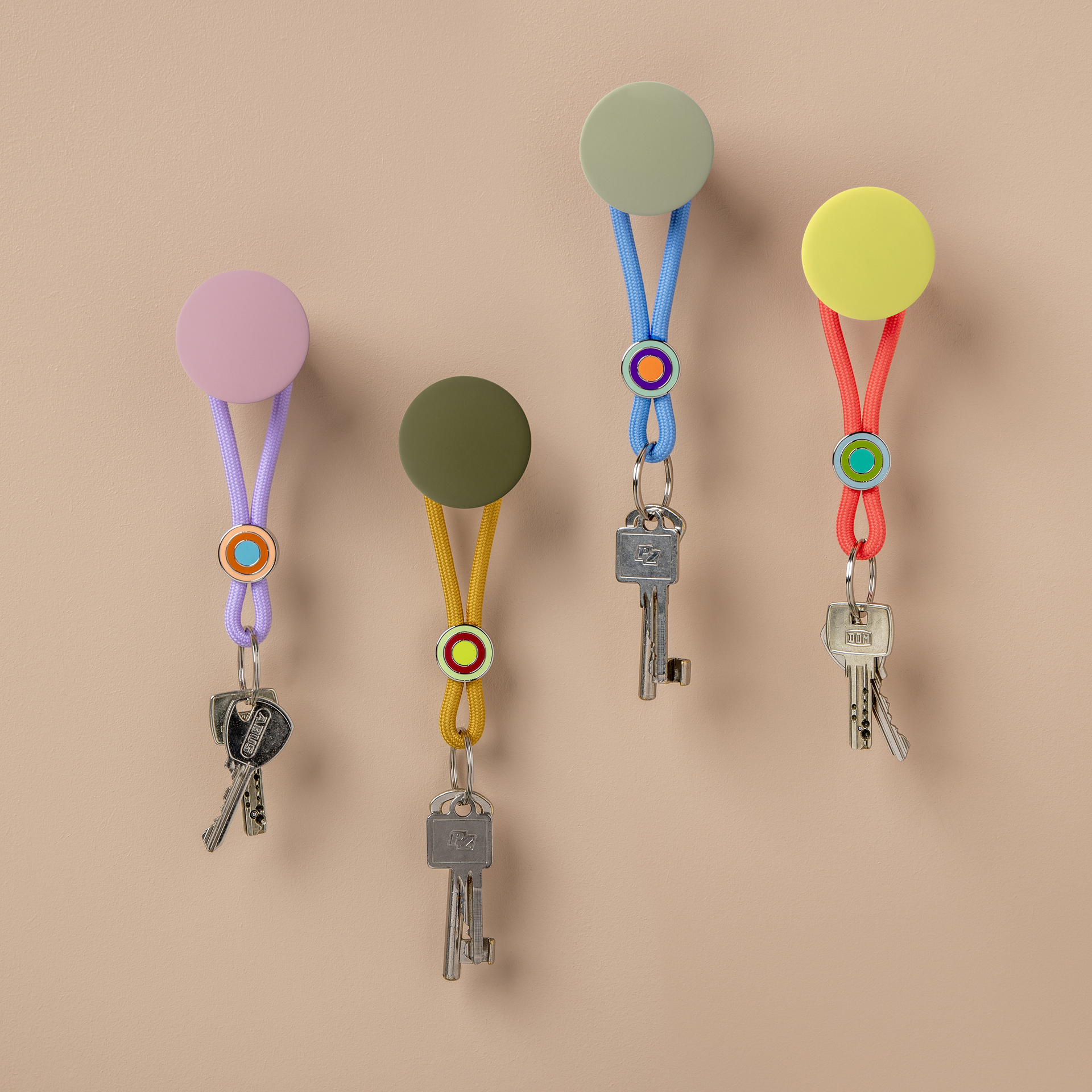 Vier farbenfrohe Schlüsselanhänger sind im Shop als Hangtags an einer Wand präsentiert. Die Anhänger haben runde Köpfe in Lila, Grün, Gelb und Gelb mit farblich passenden Kordeln. An den Kordeln hängen Schlüssel.
