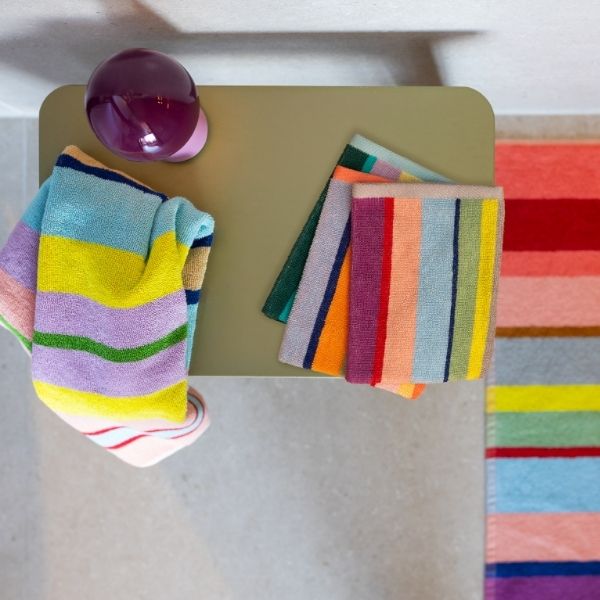 Composition colorée de serviettes rayées et d'un tapis assorti. Une grande serviette pastel est suspendue à une table vert clair, près d'une boule violette brillante. Trois serviettes pliées vives sont posées dessus.